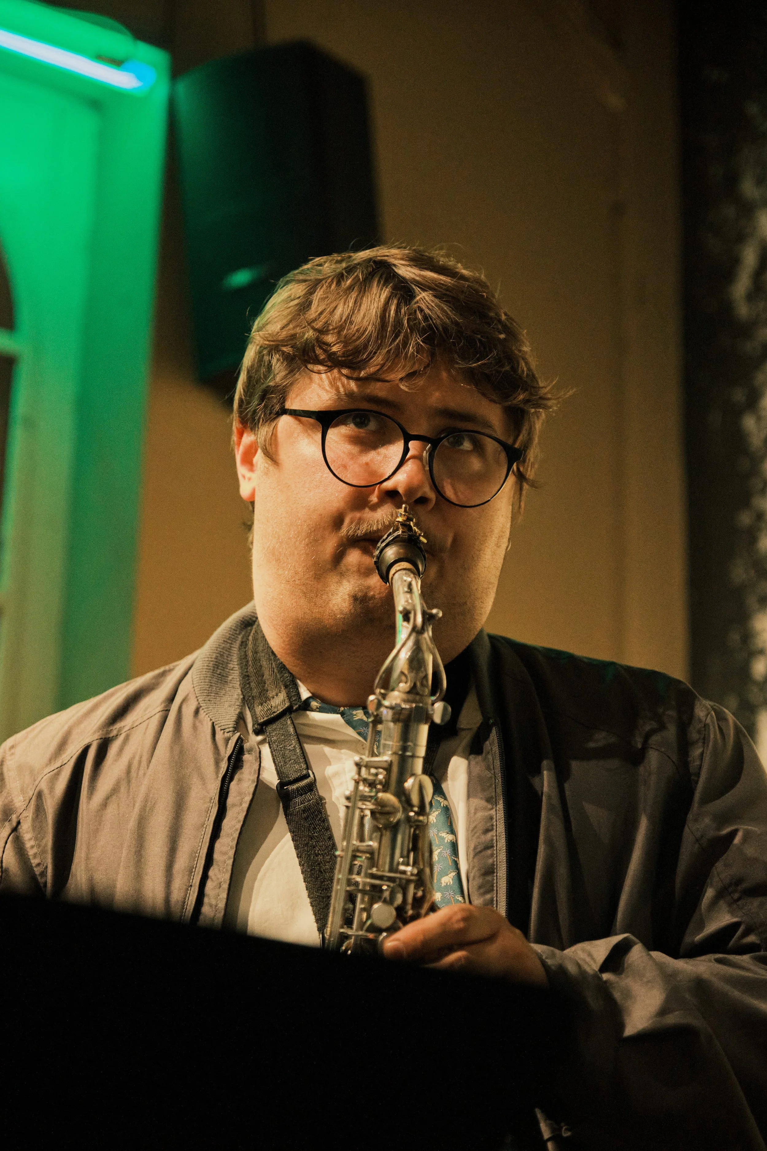 A man with glasses playing a saxophone in a dimly lit indoor setting.