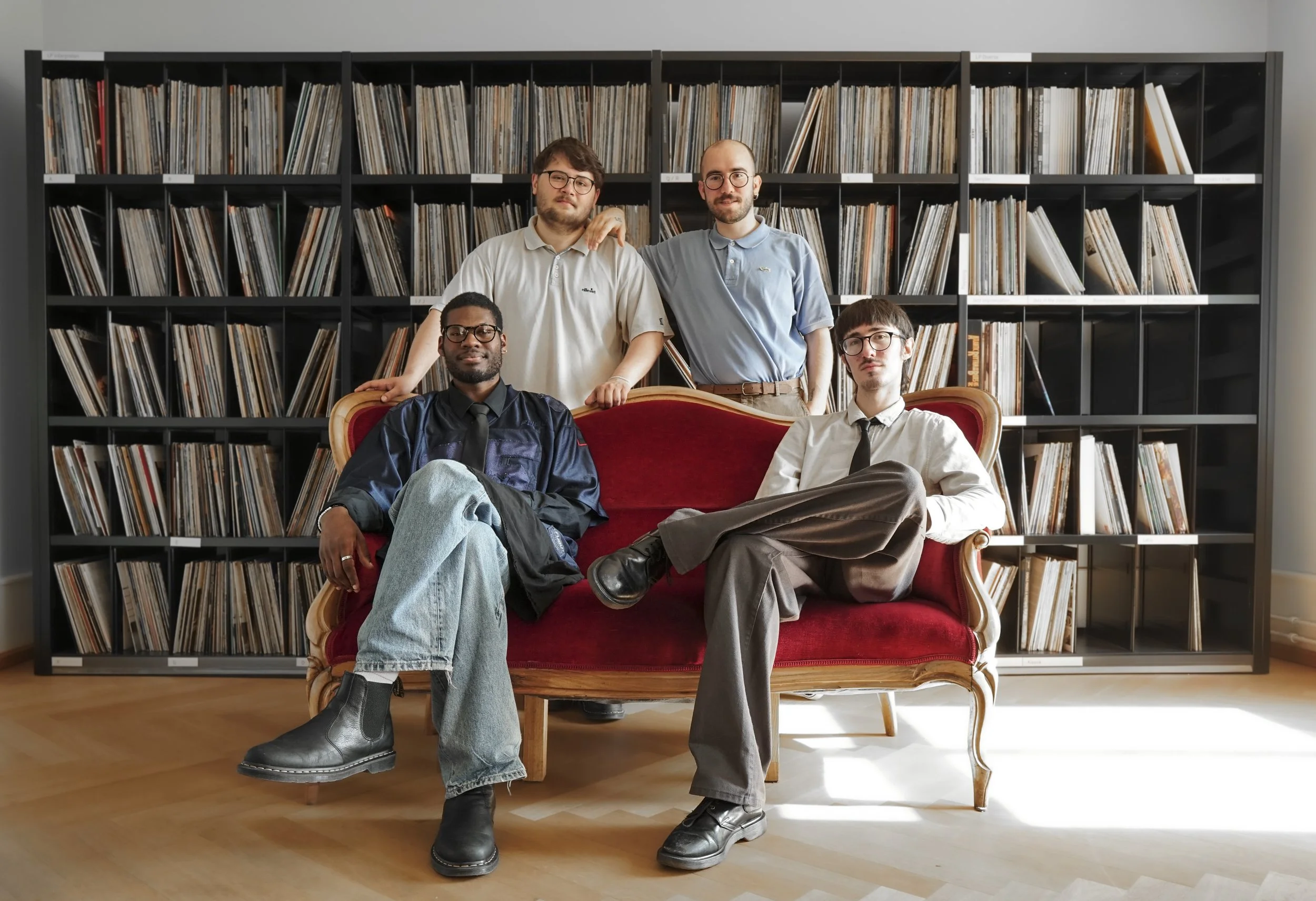 Four men in a room with a bookshelf filled with vinyl records. Two are seated on a red vintage sofa, and two are standing behind it. The setting is indoors with hardwood floors and gray walls.