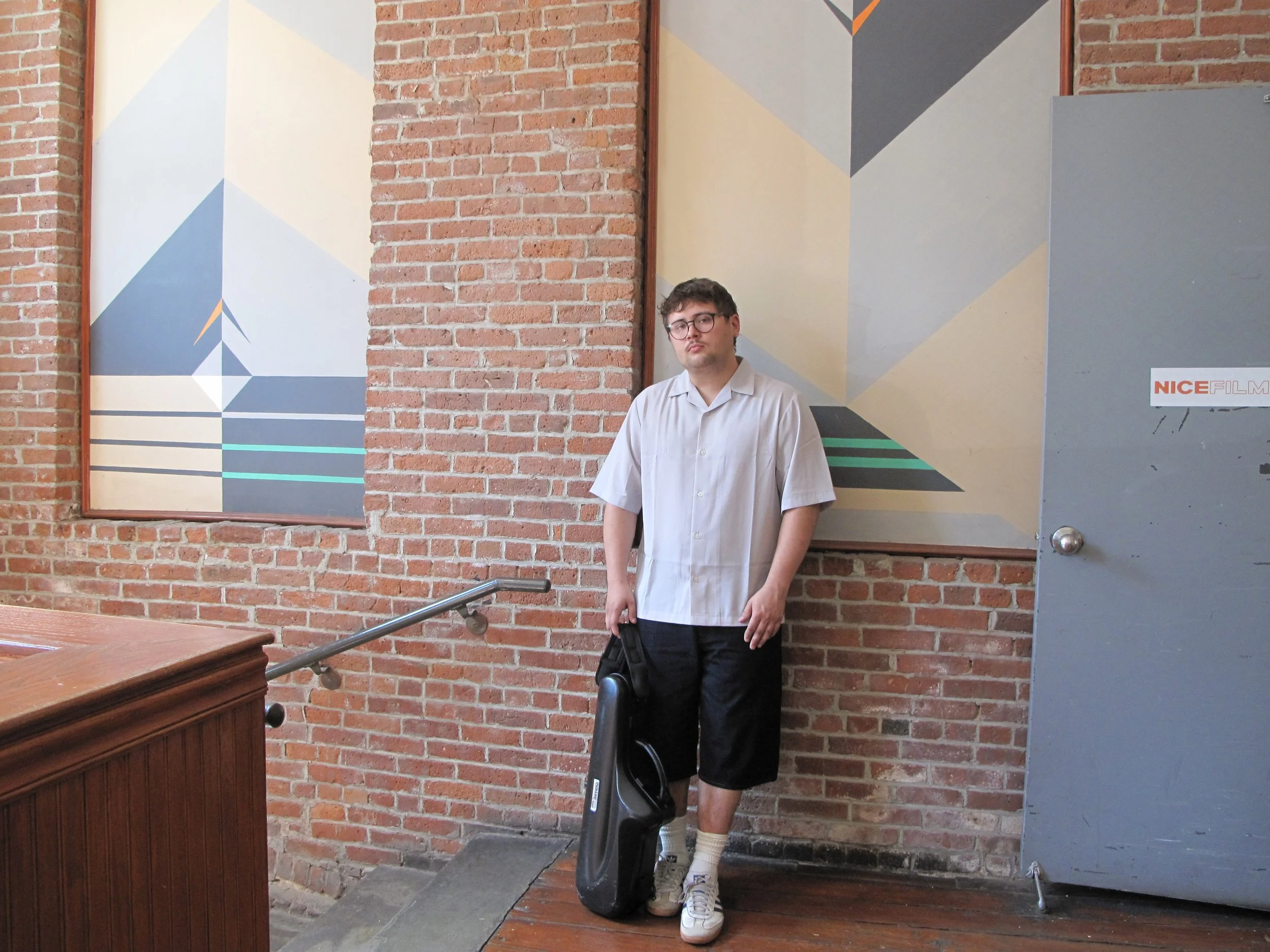 Young man in a casual shirt, shorts, white sneakers, standing against a brick wall with geometric art, holding a black bag, with a guitar case on the ground.