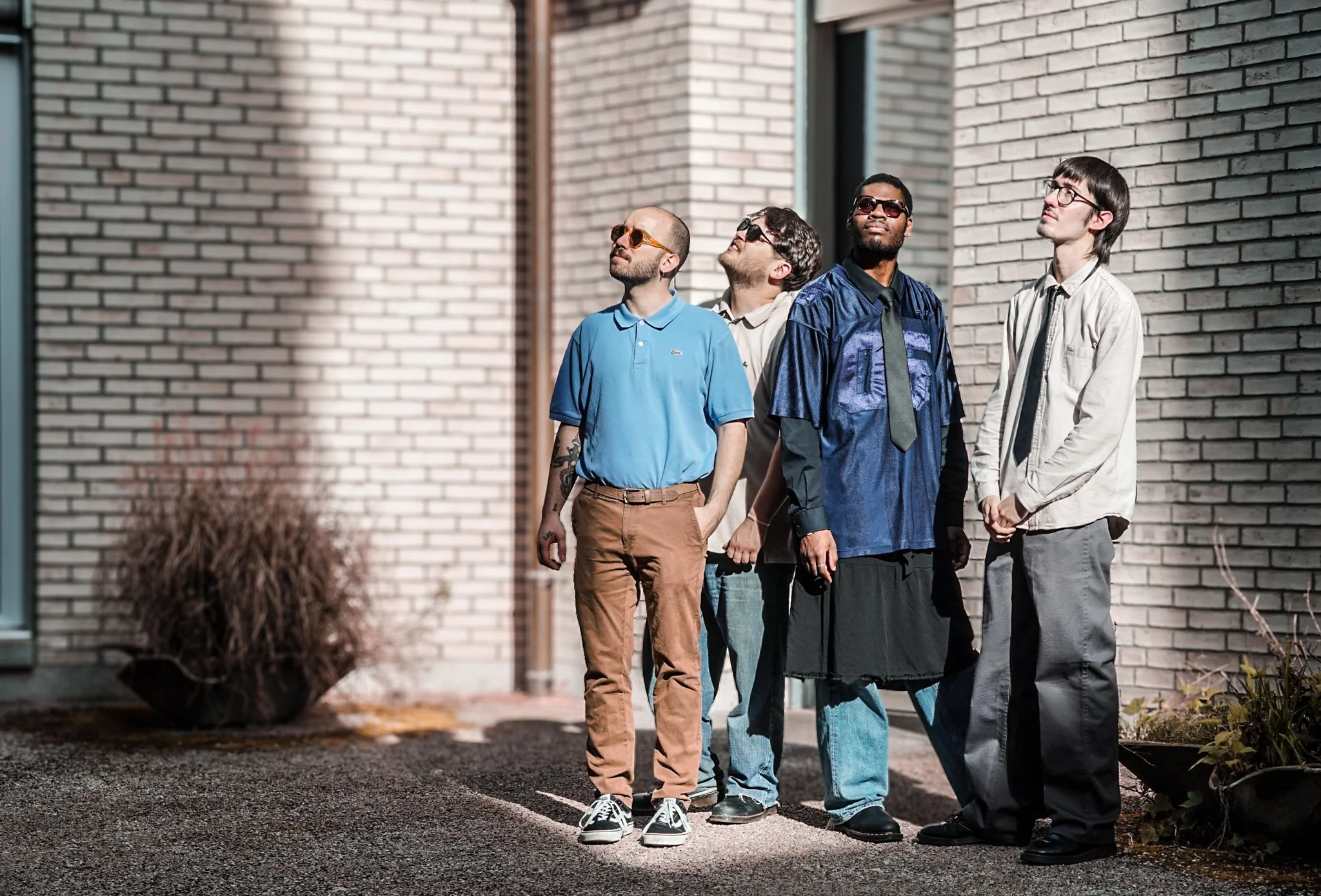 Five casually dressed men standing on a sidewalk near a brick wall, looking upwards.