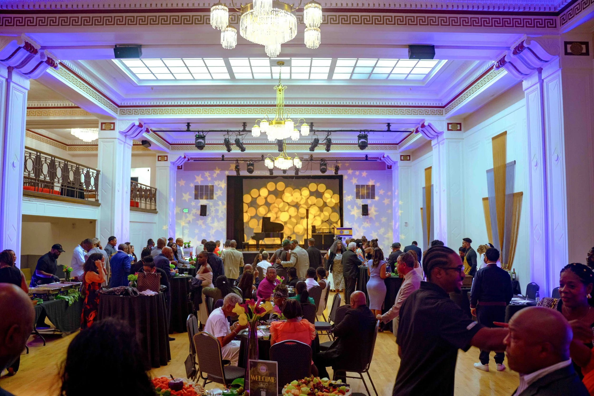 A large banquet hall filled with people attending a formal event. The room has white walls, decorative moldings, chandeliers, and a stage at the front with a grand piano and a backdrop with star and circle patterns. There are tables with green tablec