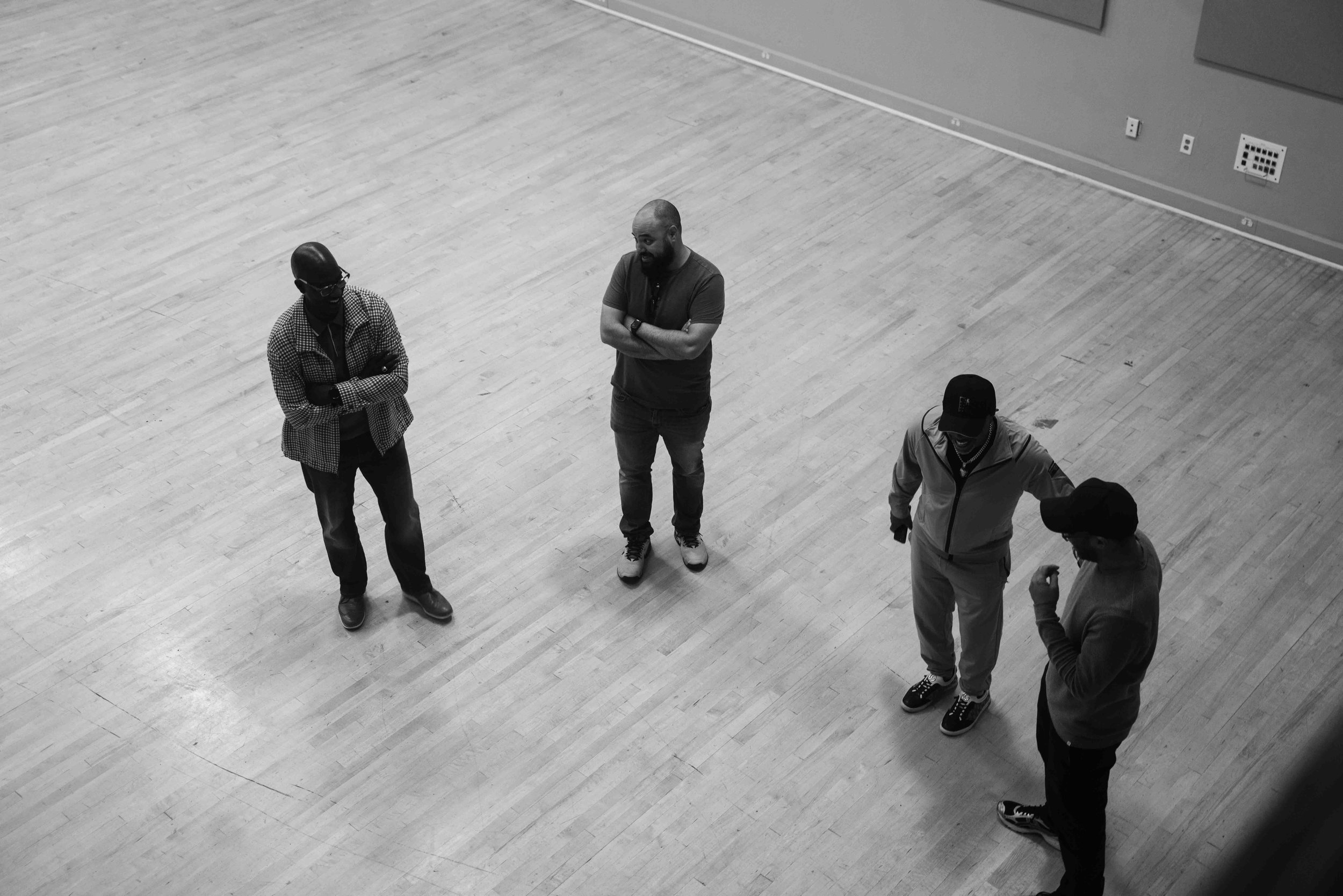 Four men standing on a wooden floor in a spacious room, engaged in conversation.
