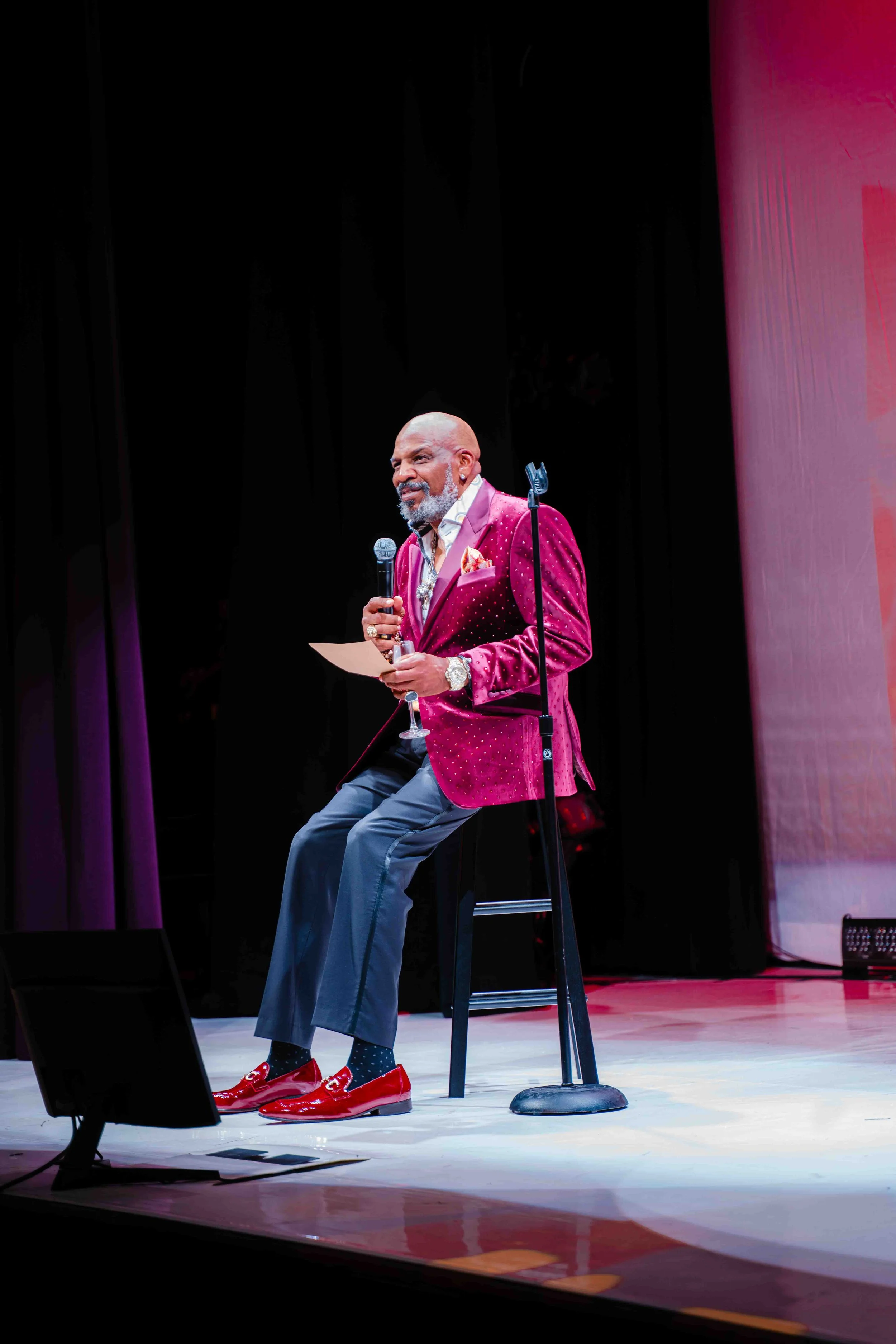 A man in a red velvet suit jacket, gray trousers, and shiny red shoes is sitting on a tall stool on stage, holding a microphone and a piece of paper, while performing or speaking at an event. The stage has a dark background with a lit area in front o