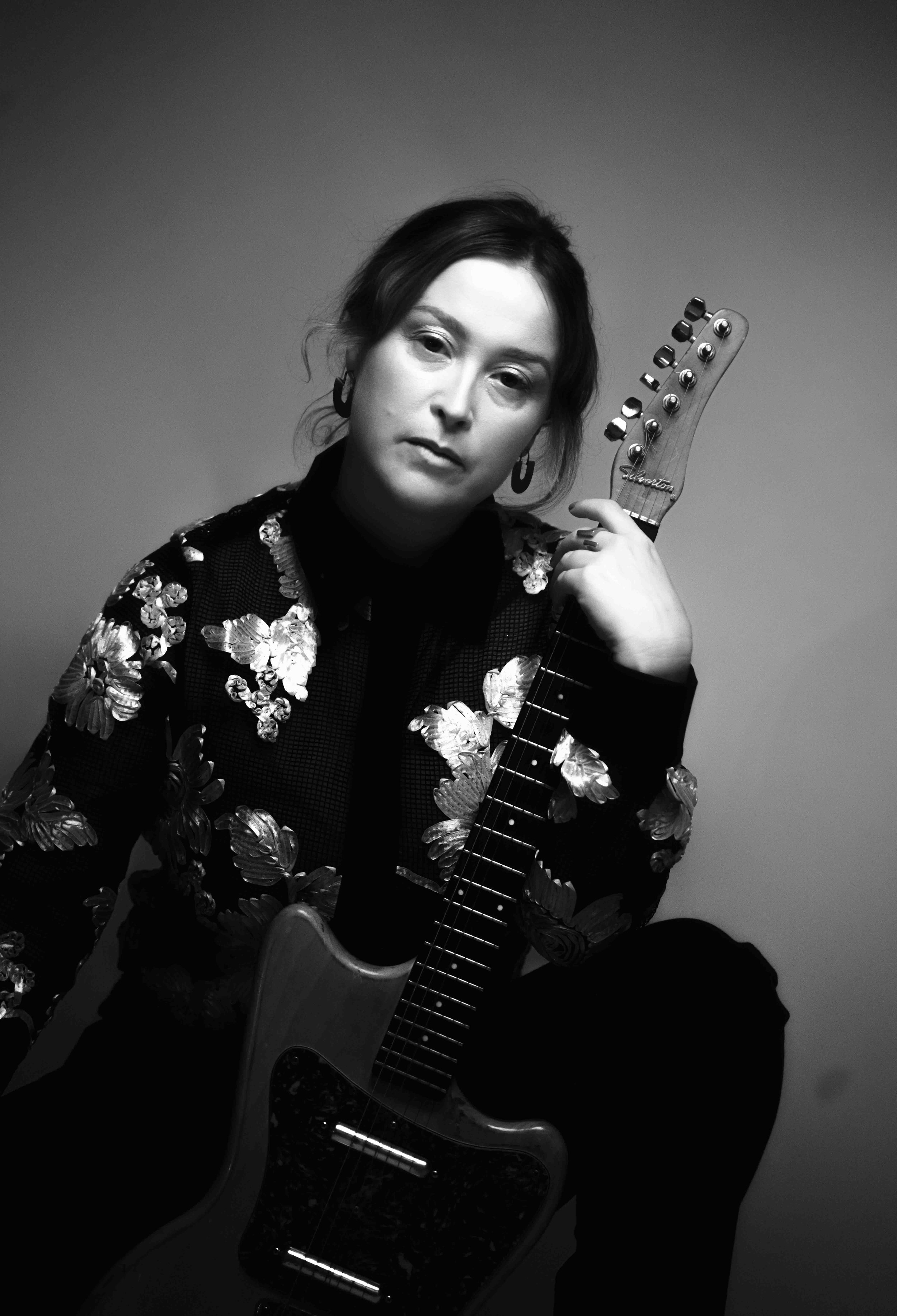 Black and white photo of a woman with dark hair holding an electric guitar, wearing a floral embroidered jacket and earrings, against a plain background.