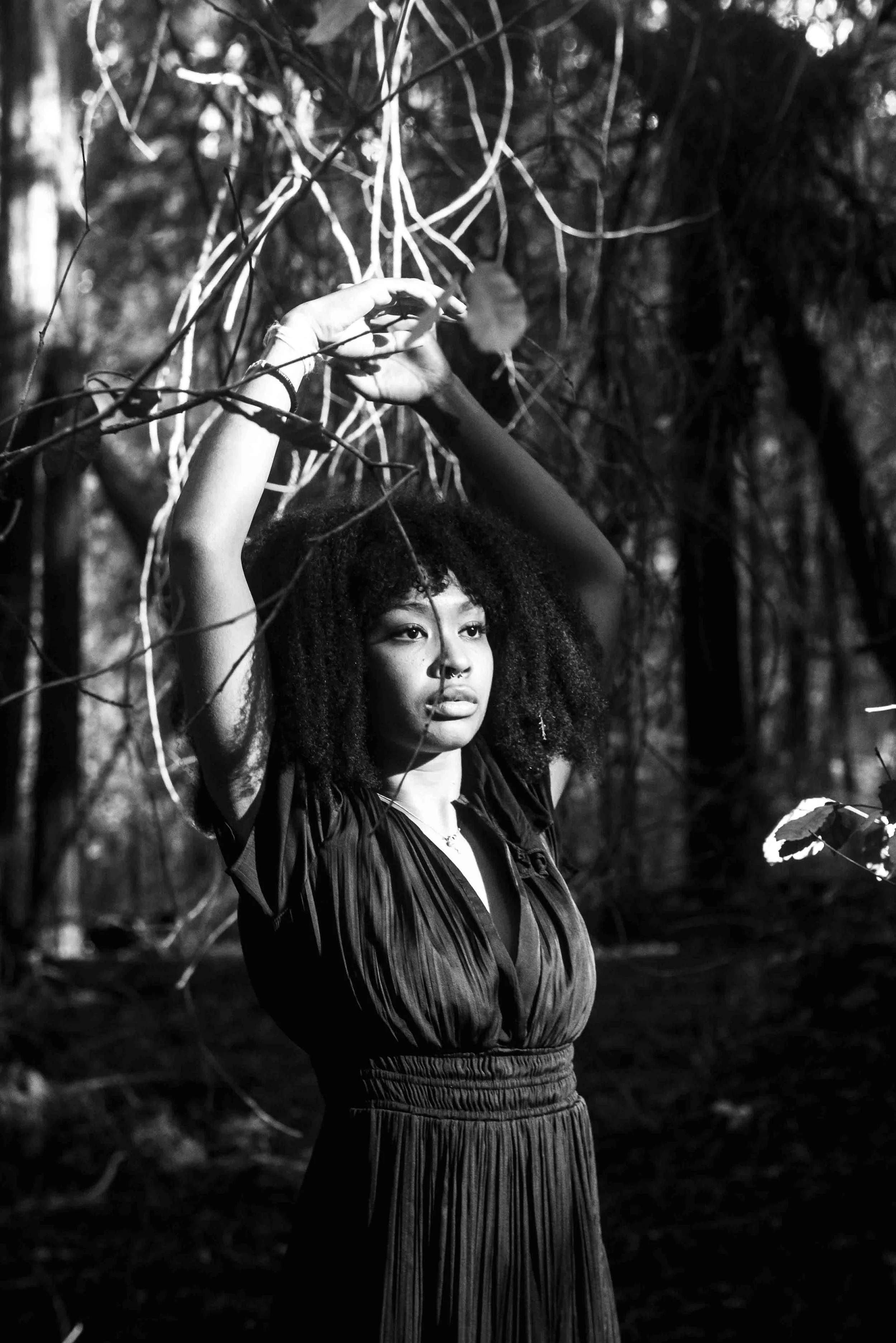 A young woman with curly hair standing in a wooded area, raising her arms to hold onto tree branches.