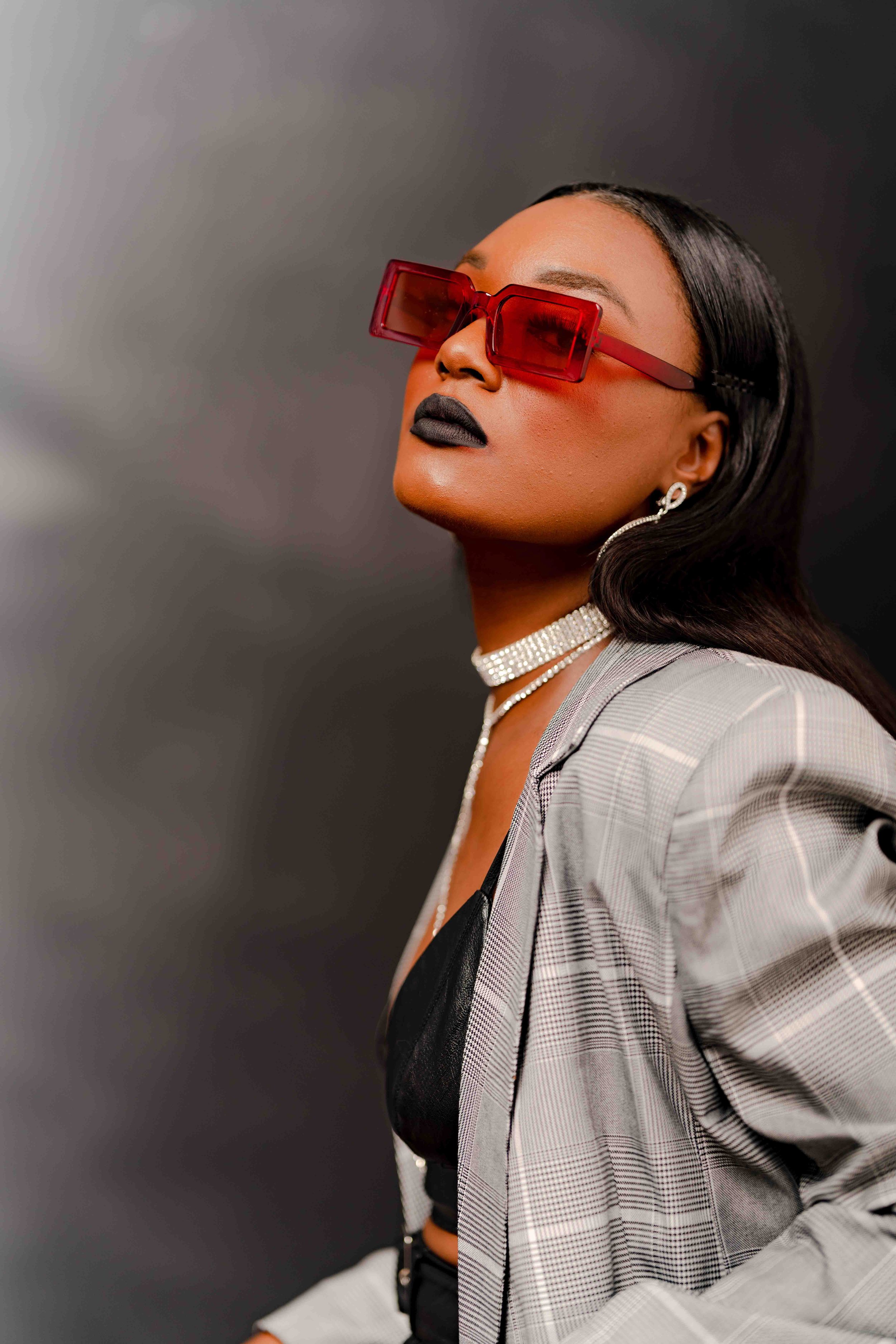 A woman wearing large red sunglasses, black lipstick, silver jewelry, and a gray plaid blazer over a black top, posing against a dark background.