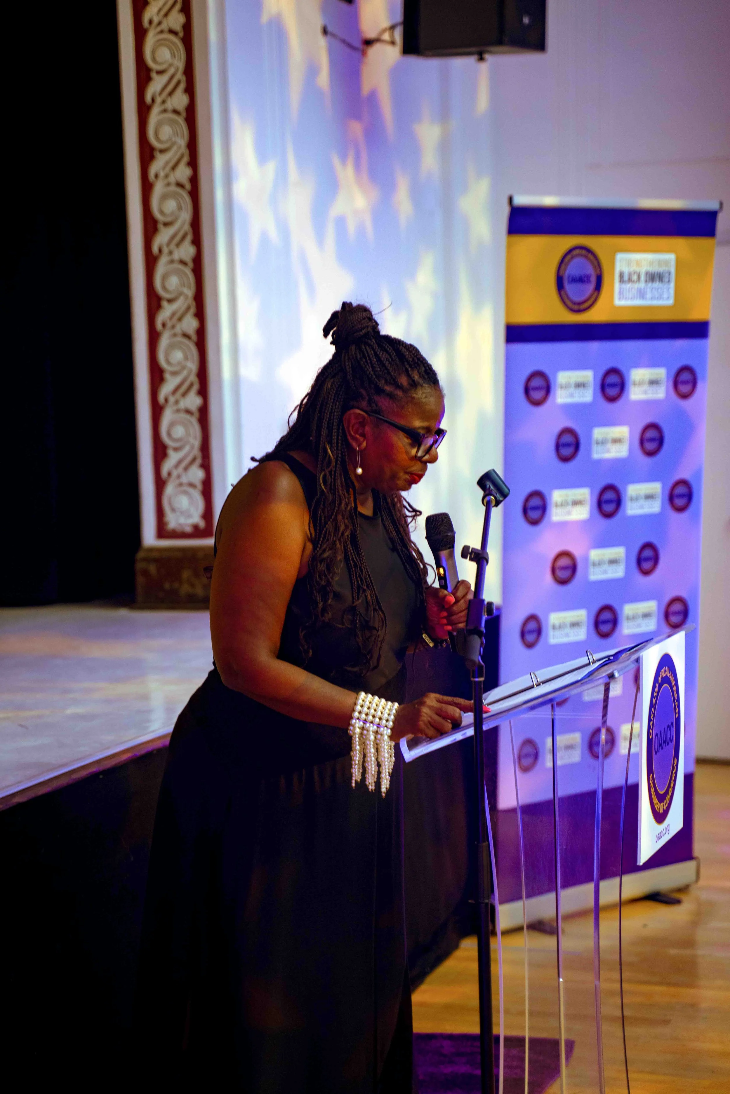 A woman with long dreadlocks and glasses standing at a podium, reading or speaking into a microphone at an event with a backdrop and a banner with logos.