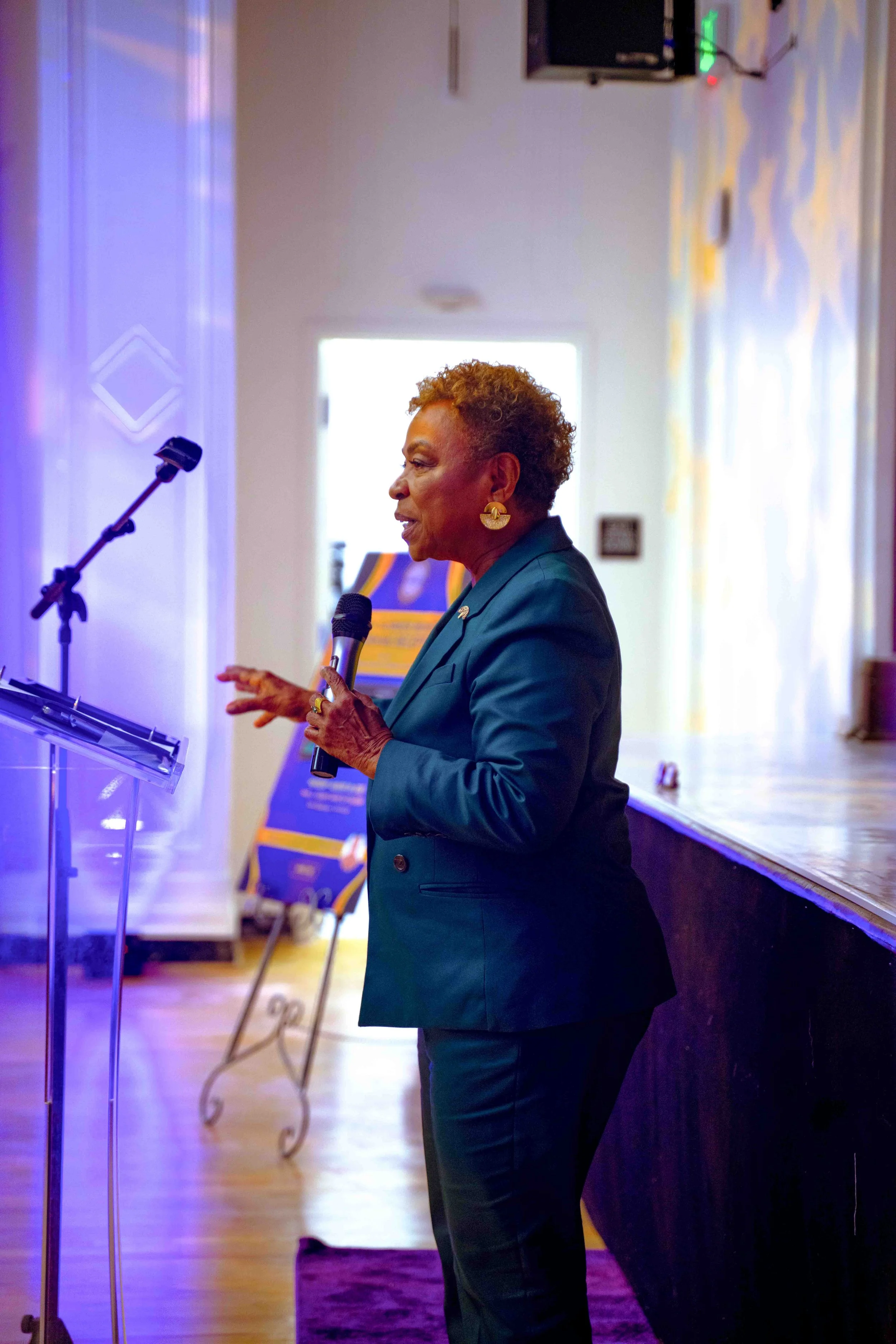 An older woman with short curly hair giving a speech at a podium, holding a microphone, in a room with a wooden floor and decorated walls.