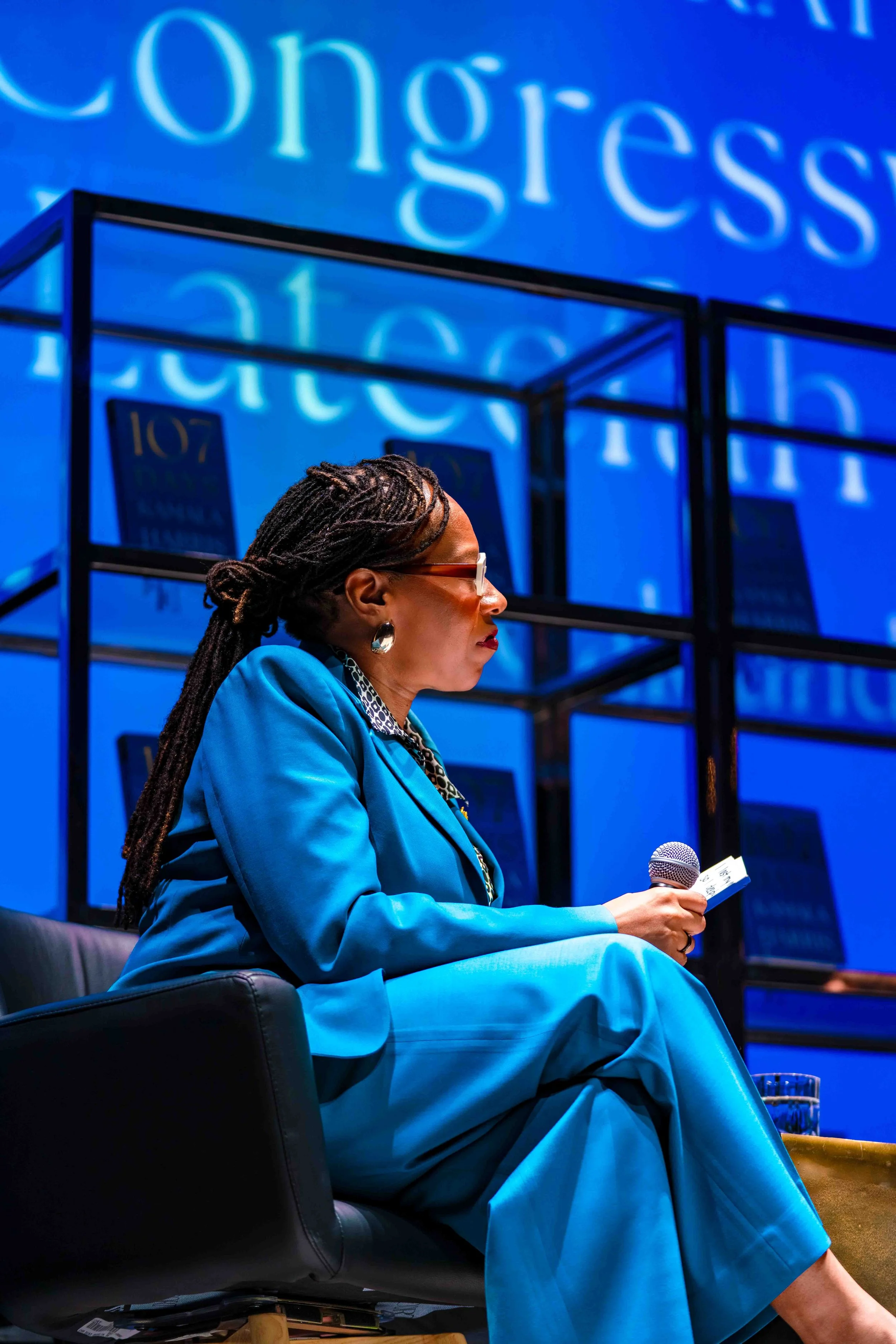 A woman sitting on a black chair, holding a microphone and a small piece of paper, wearing a blue suit and glasses, with shelves behind her and a large blue screen displaying the word 'Congress' in the background.