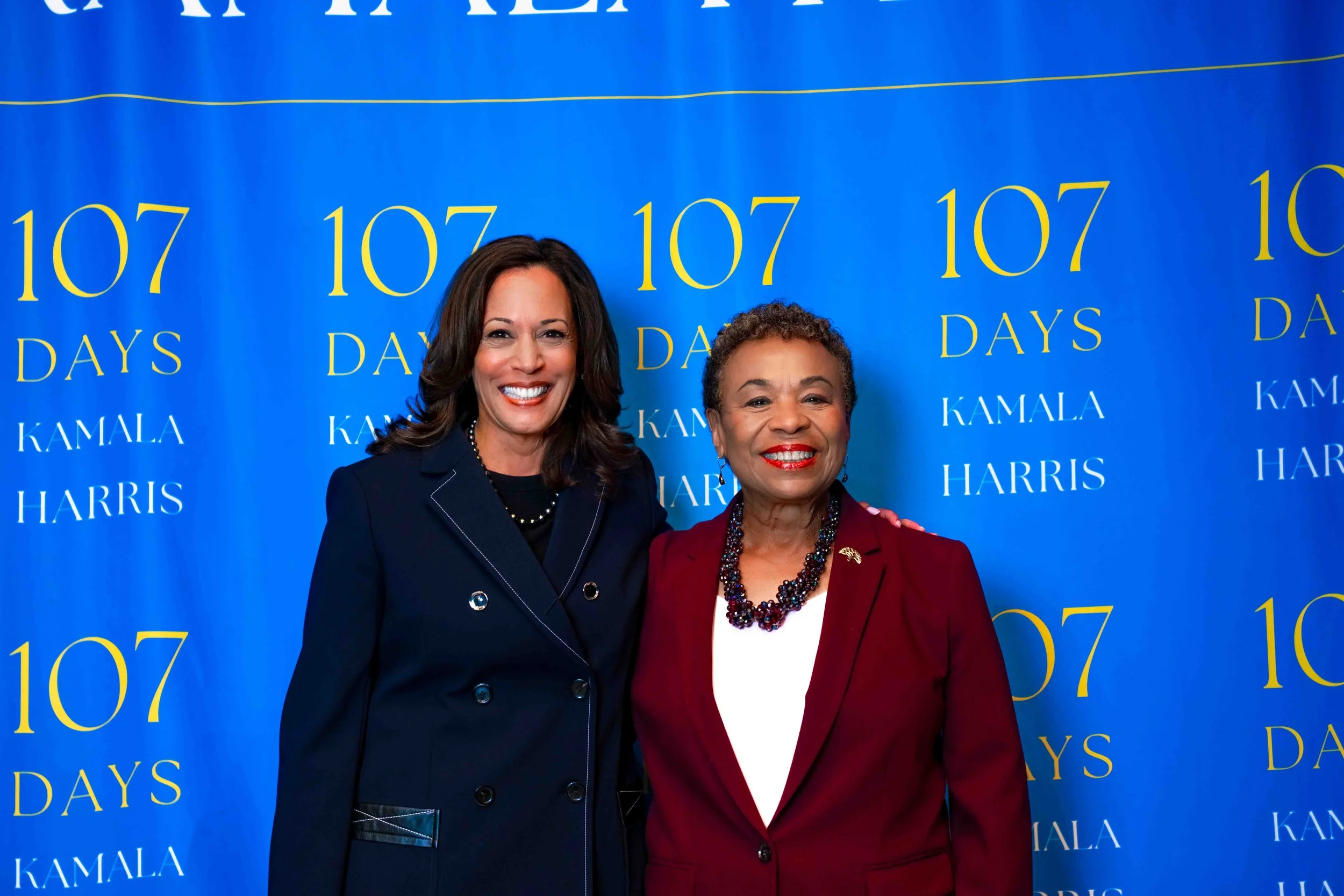 Two women standing together in front of a blue background with yellow and white text reading "107 Days Kamala Harris." Both women are smiling and wearing formal suits, with the woman on the left in a dark navy blazer and the woman on the right in a b