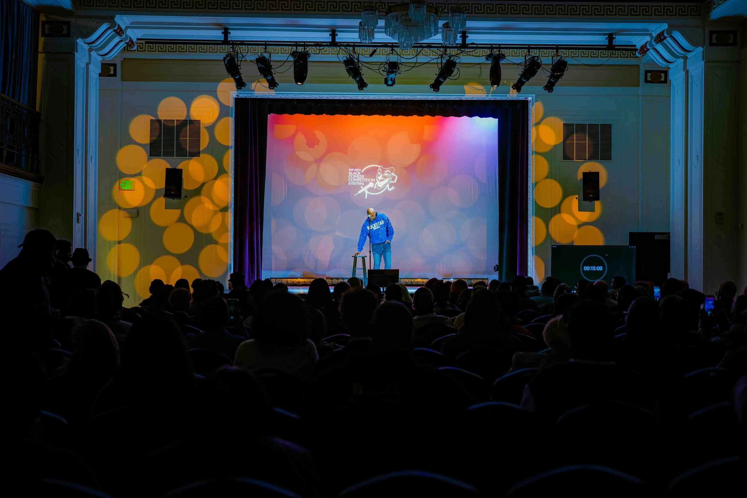 A comedian performing on stage at a comedy festival in a theater, with an audience seated in front. The stage features colorful lighting, with a backdrop displaying the event's logo.