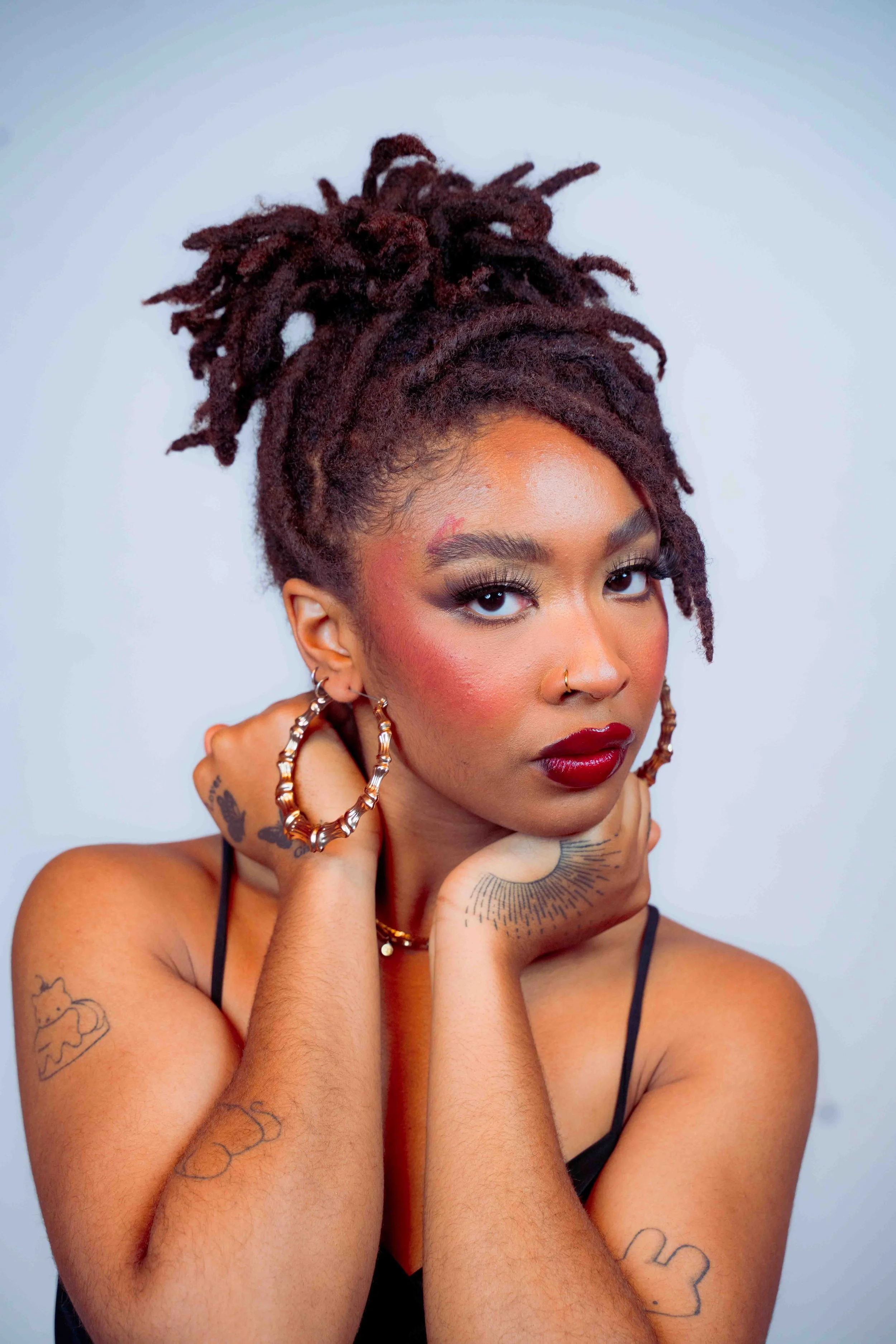 A woman with dark red dreadlocks styled in an updo, wearing bold makeup including dark lipstick and eye makeup, and dressed in a black spaghetti strap top. She has tattoos on her arms and face, large hoop earrings, and a nose ring, posing with her ha