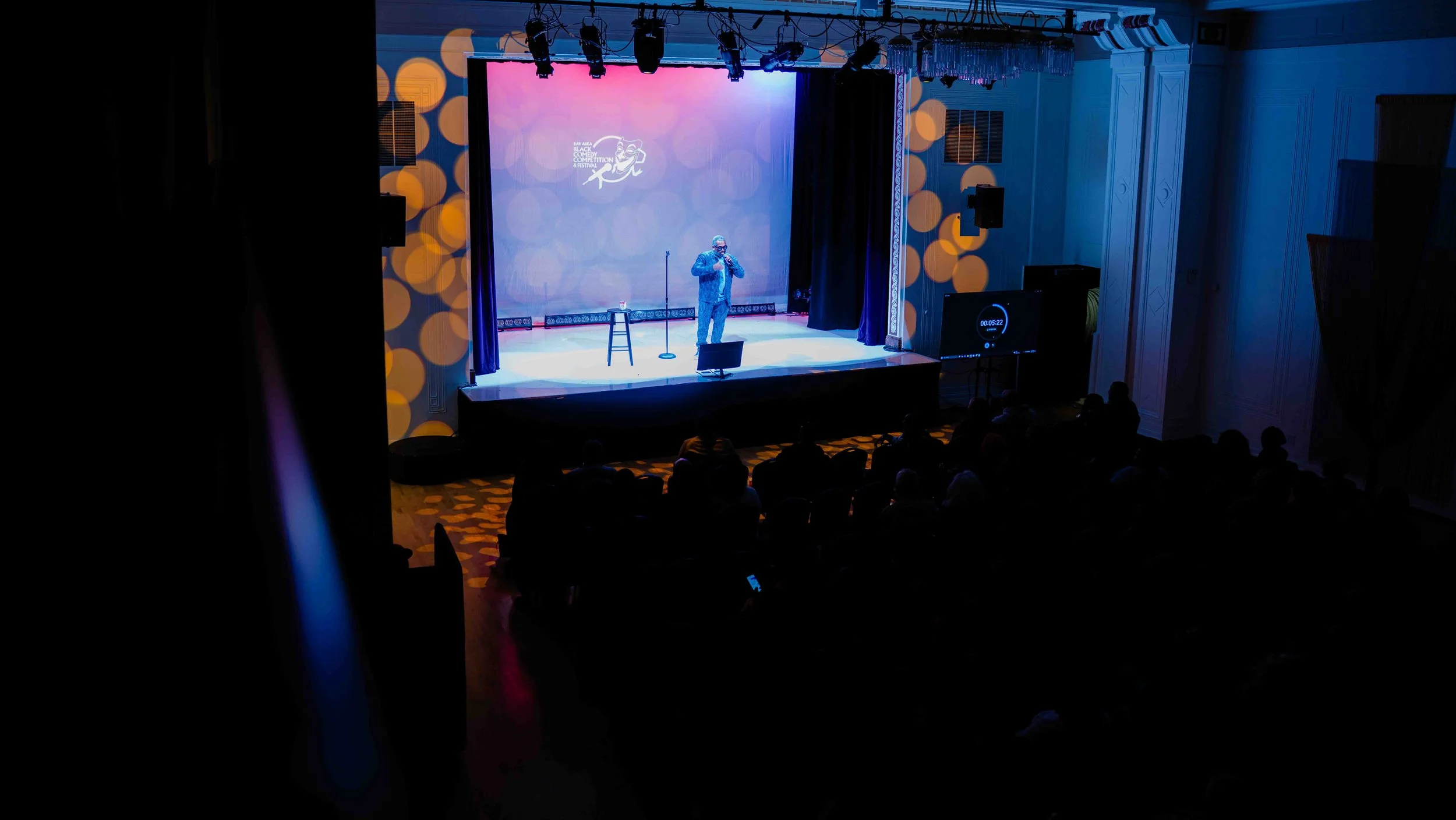 A performer on stage at a comedy club or event, with a microphone and stool, illuminated by stage lighting, and an audience watching in the dark.