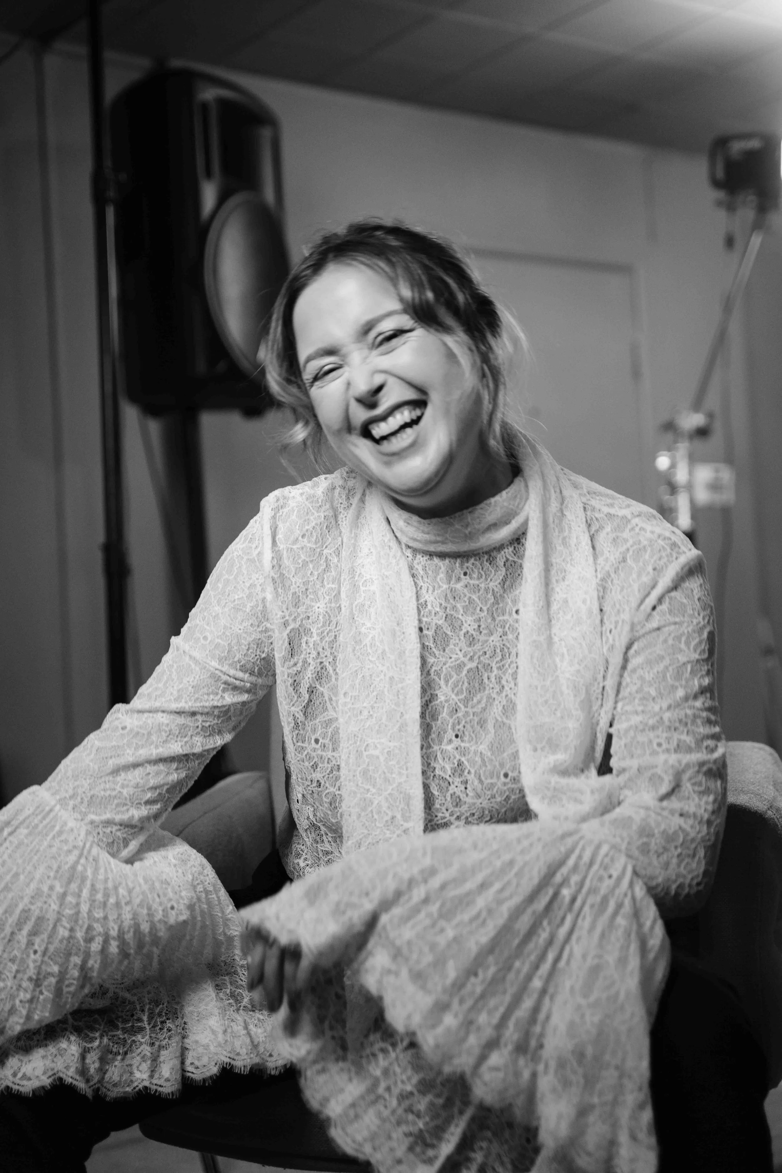 A woman in a lace dress sitting and laughing in a room with large speakers.