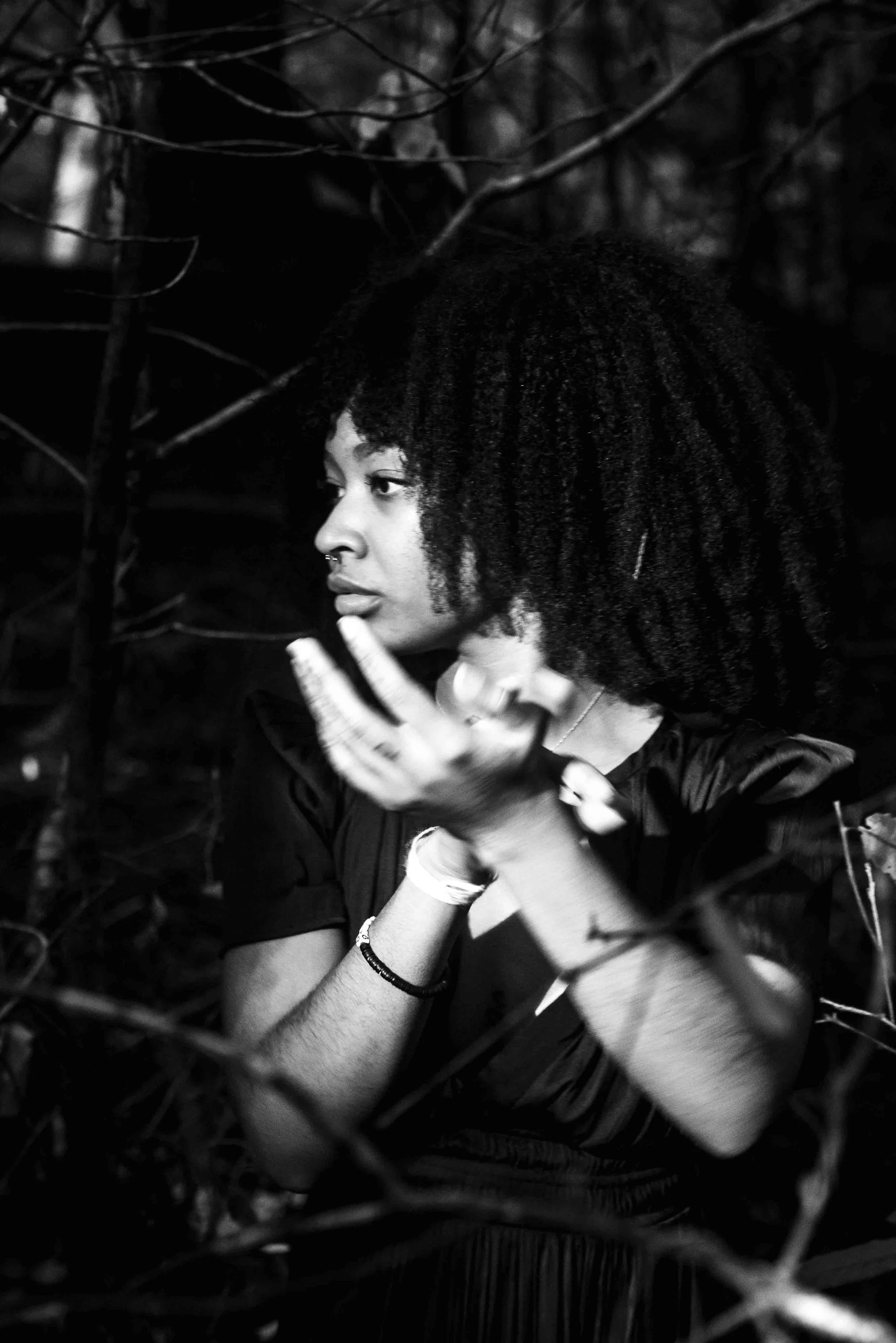 A woman with natural curly hair looking to the side, sitting among branches and leaves, in a black and white photograph.