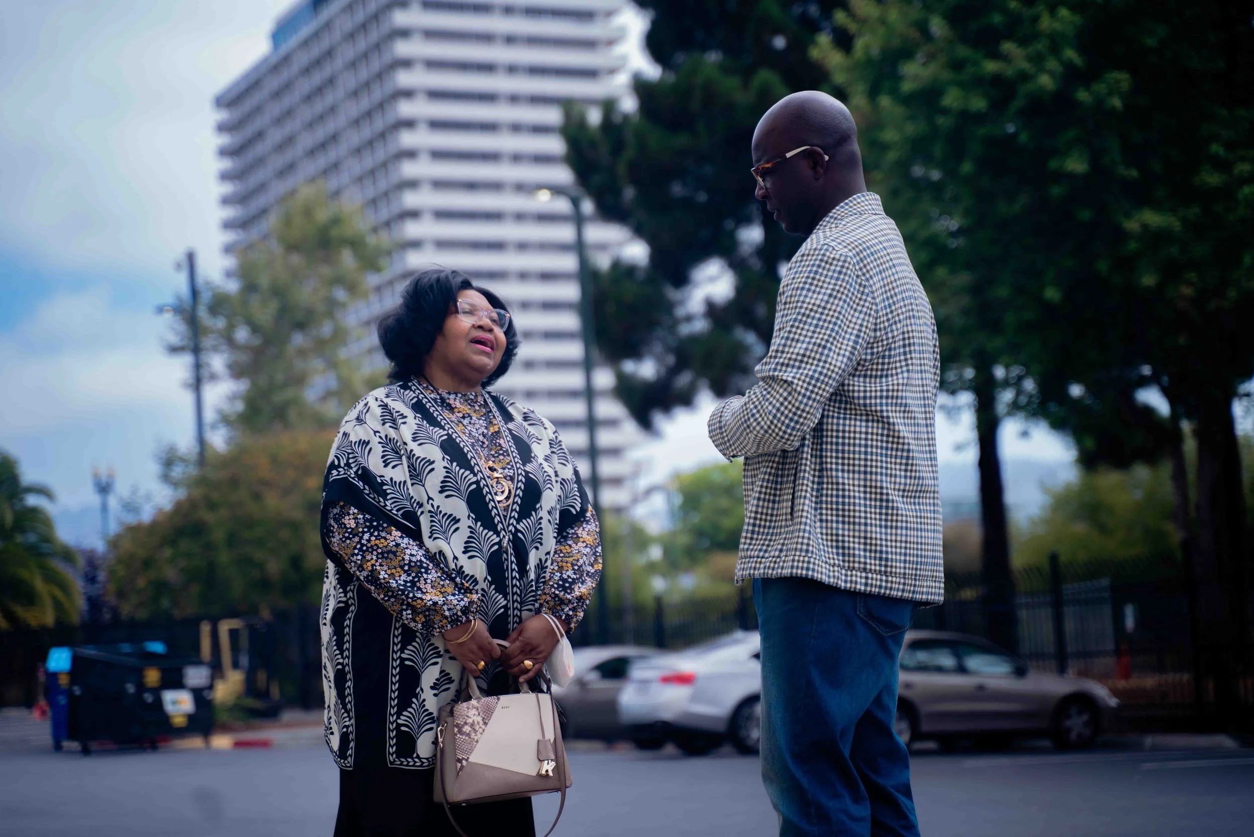 An elderly woman and a man having a conversation outdoors in an urban area, with a tall building and trees in the background.