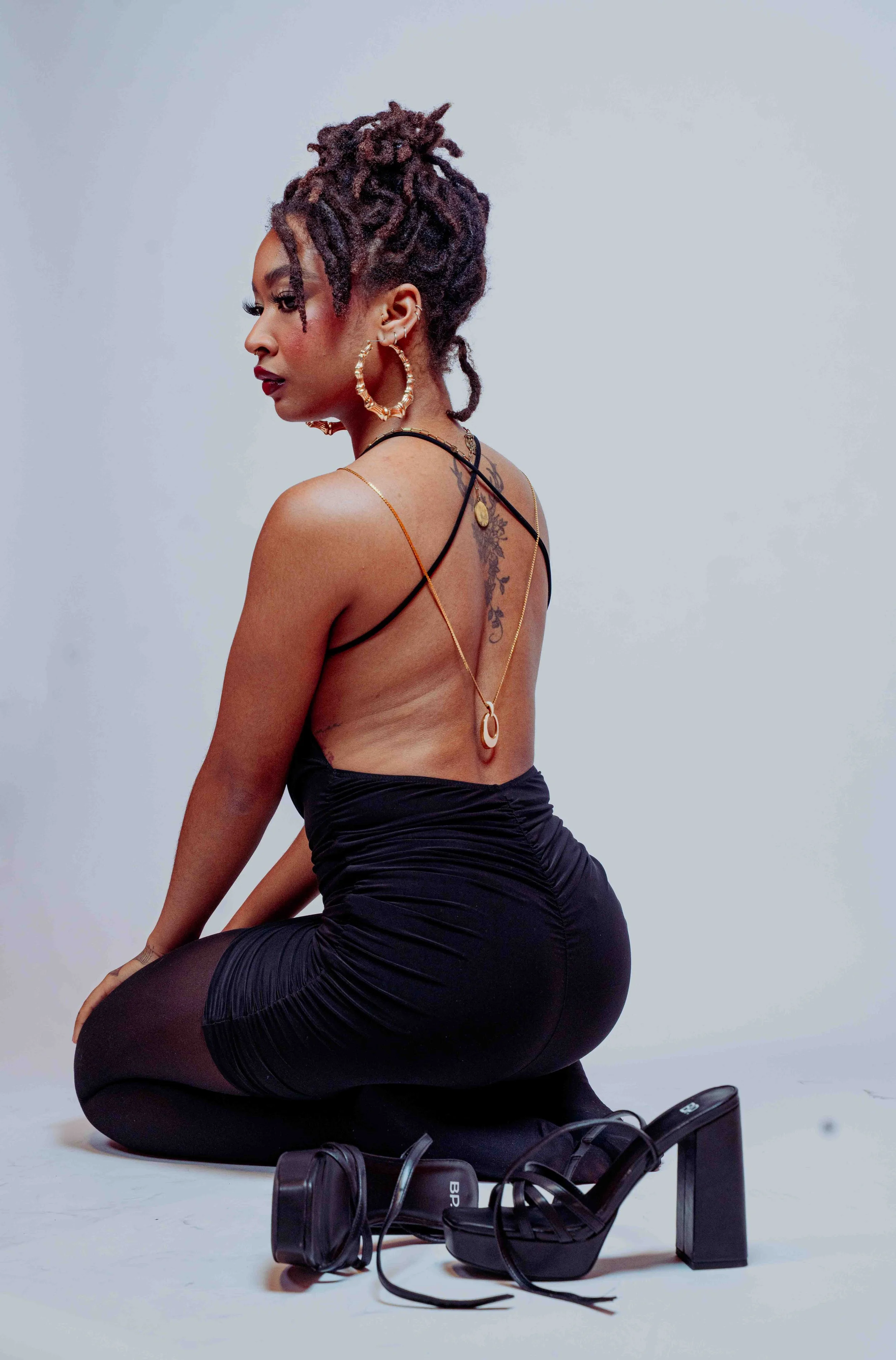 A woman with short, curly hair kneeling on the floor, wearing a black dress with open back and black high heels, with jewelry and a tattoo on her back, against a light gray background.