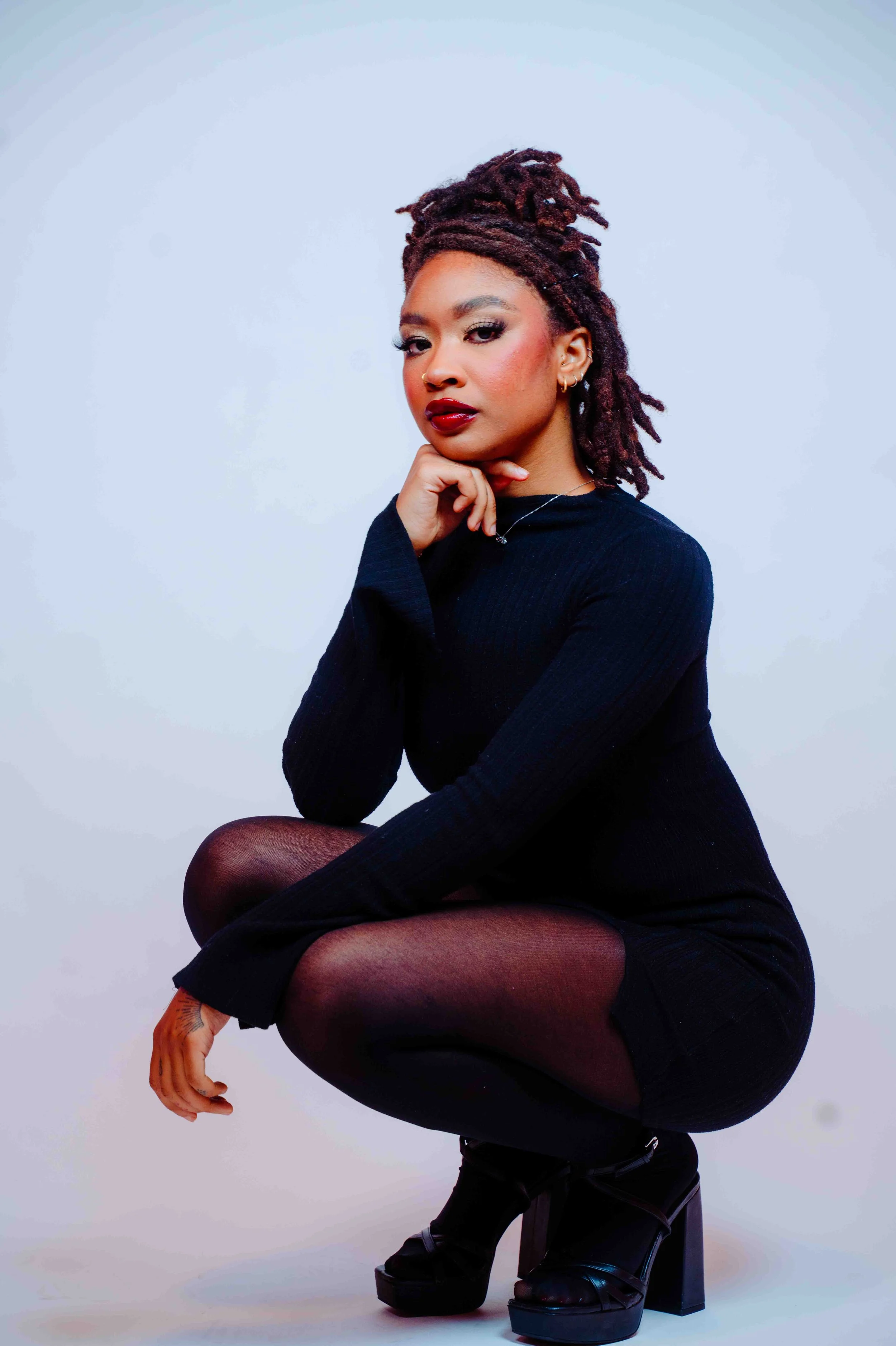 Portrait of a woman with dreadlocks, wearing a black long-sleeve dress, sitting with one knee up, against a light background.