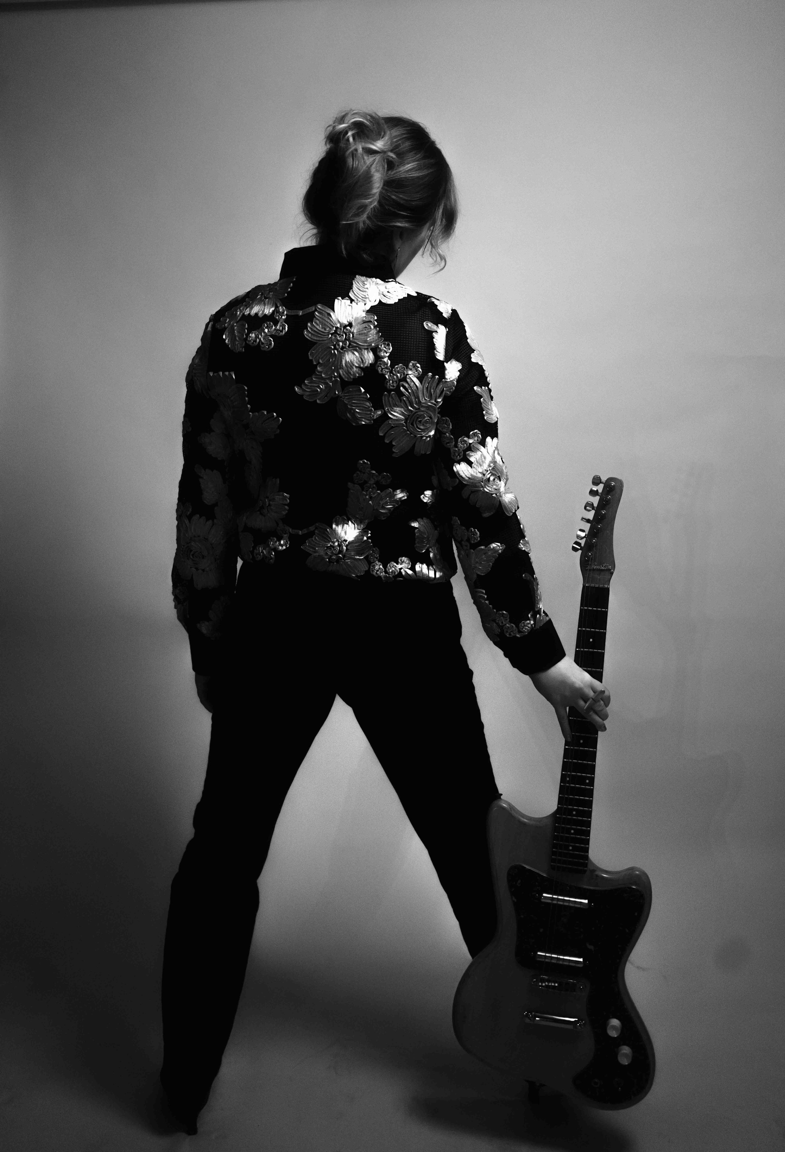 A person with short hair standing with their back facing the camera, holding an electric guitar by their side, wearing a decorated jacket, in front of a plain background.