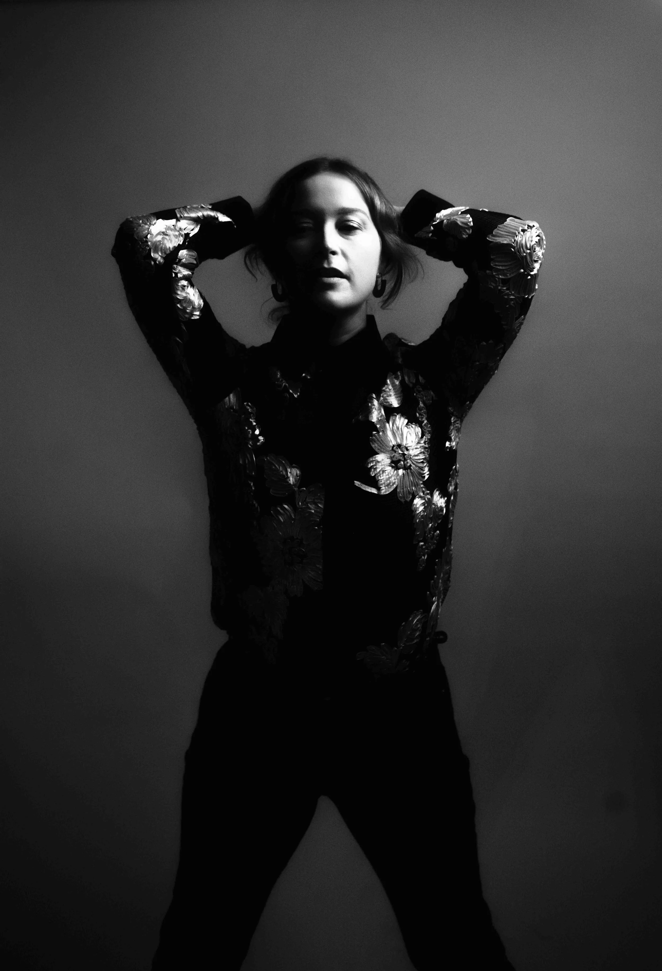 Black and white photo of a woman with arms raised behind her head, wearing a floral-patterned top, standing against a plain background.