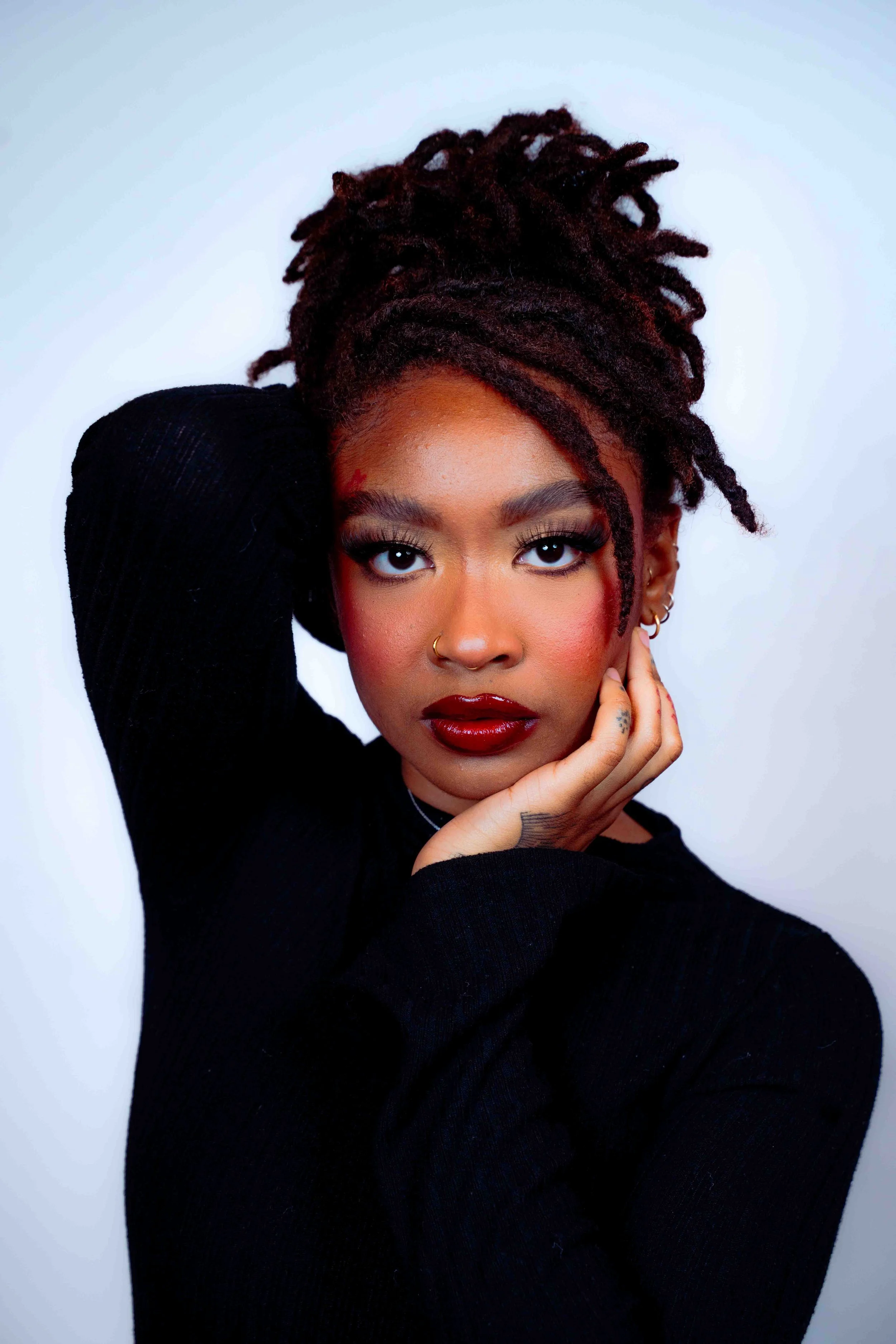 A woman with brown dreadlocks styled in an updo, wearing a black top, bold red lipstick, and gold jewelry, posing with her hand near her face against a plain light background.