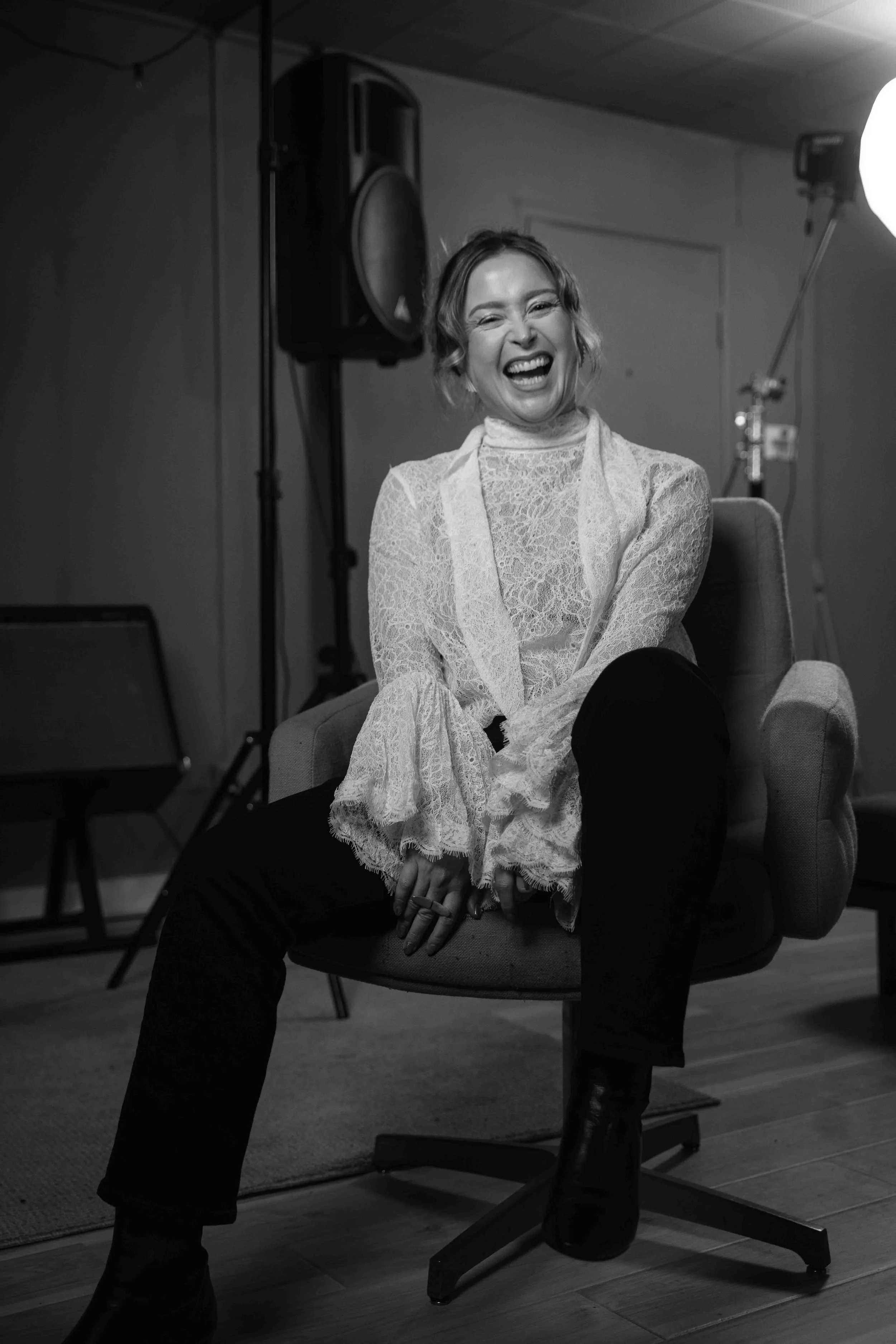 A woman sitting in a chair, laughing, in a studio setting with lighting equipment and speakers visible in the background.