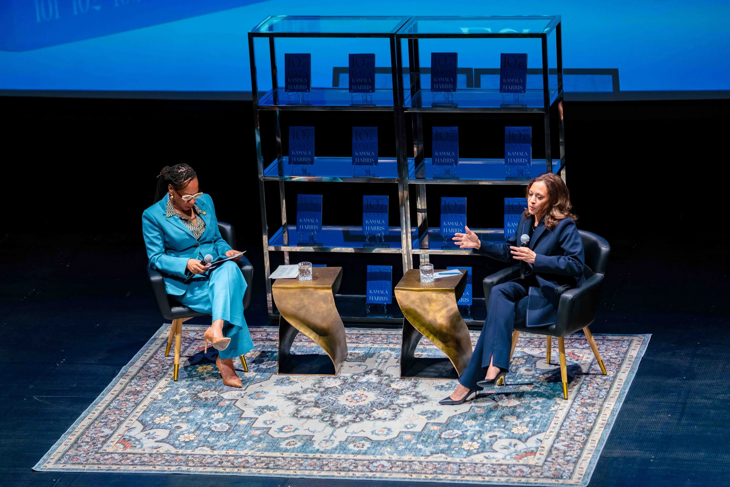 Two women seated on stage during a book discussion, with a display of Kamala Harris's book titled '107 Days' behind them. One woman is wearing a blue suit, and the other is in a dark suit, both holding microphones. There are small tables with glasses