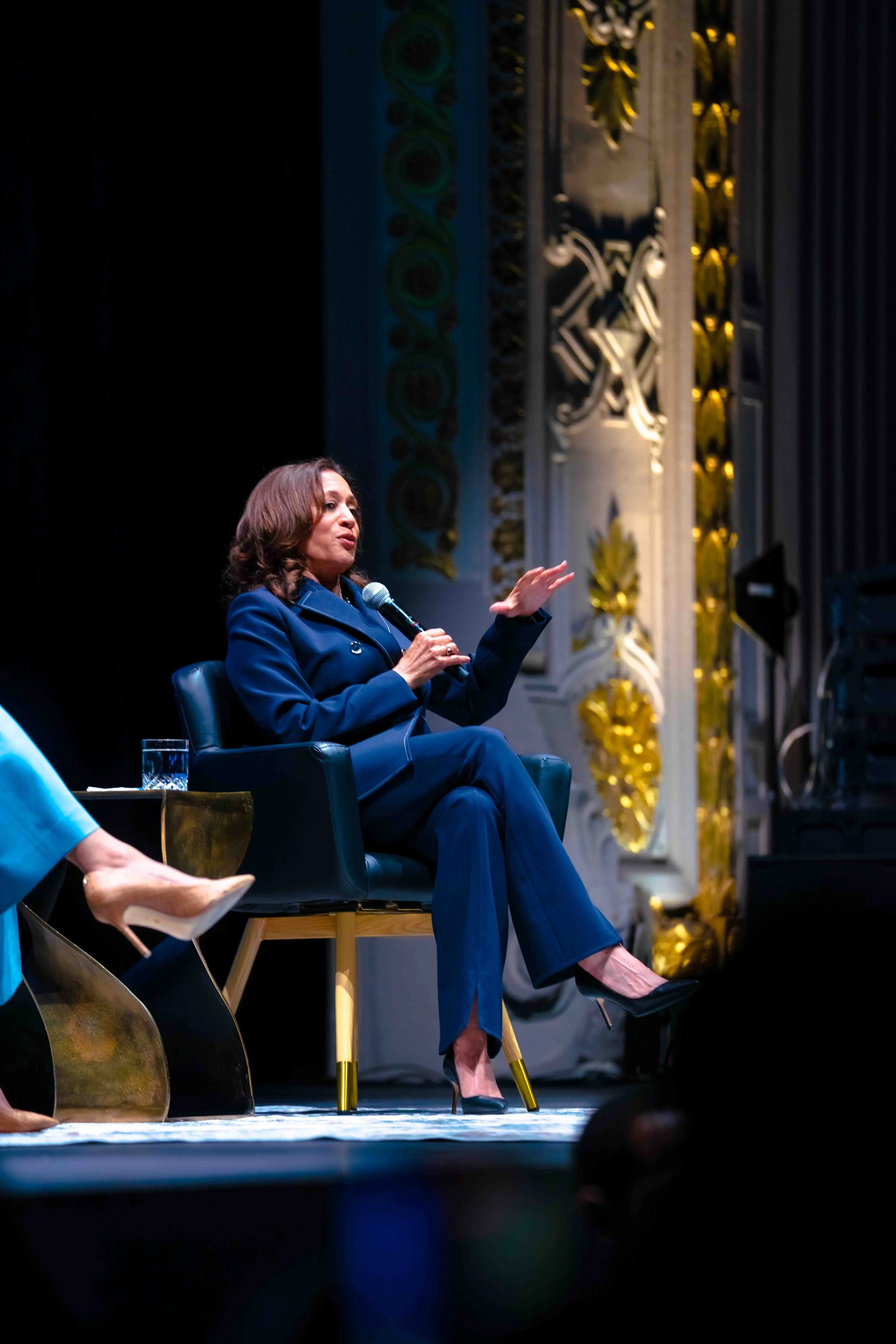 A woman in a dark blue suit is sitting on a black armchair on a stage, holding a microphone and gesturing with her left hand. She has shoulder-length brown hair and is wearing high heels. There are ornate gold and white decorative elements behind her
