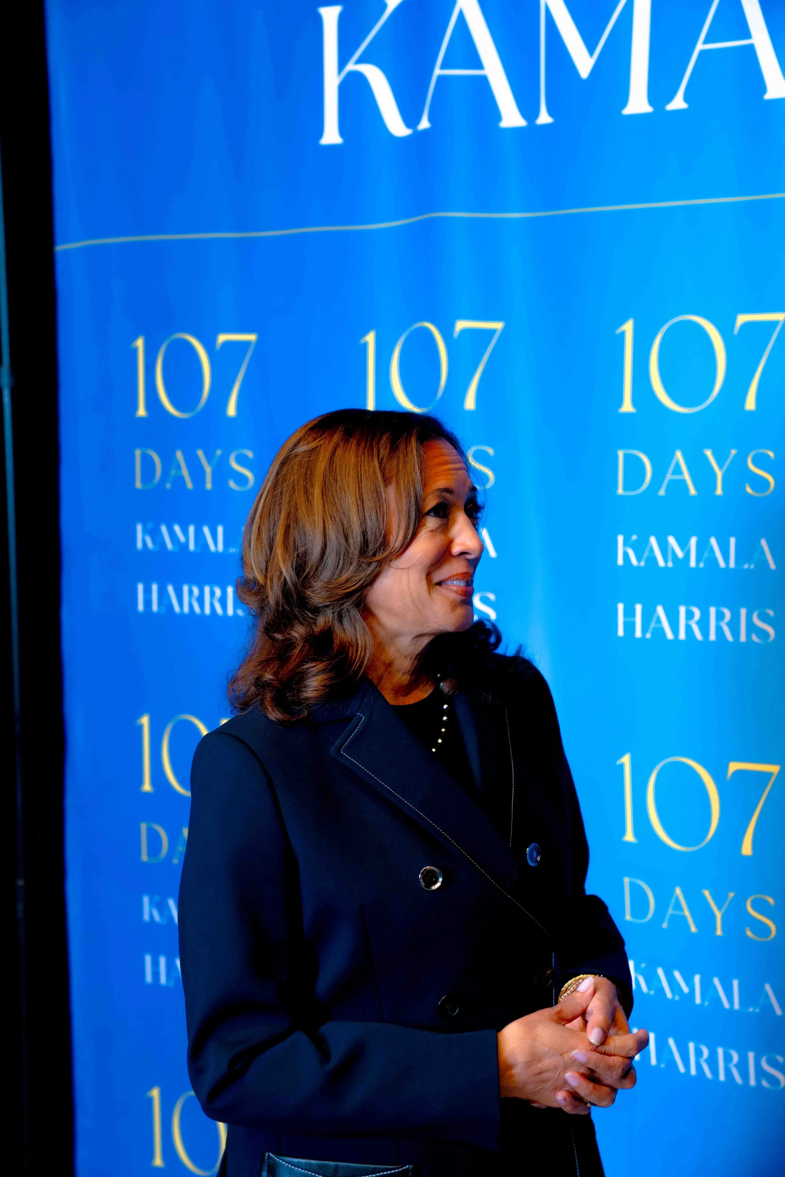A woman with shoulder-length brown hair and a dark blazer, standing in front of a blue backdrop with the text '107 Days Kamala Harris' repeated multiple times.