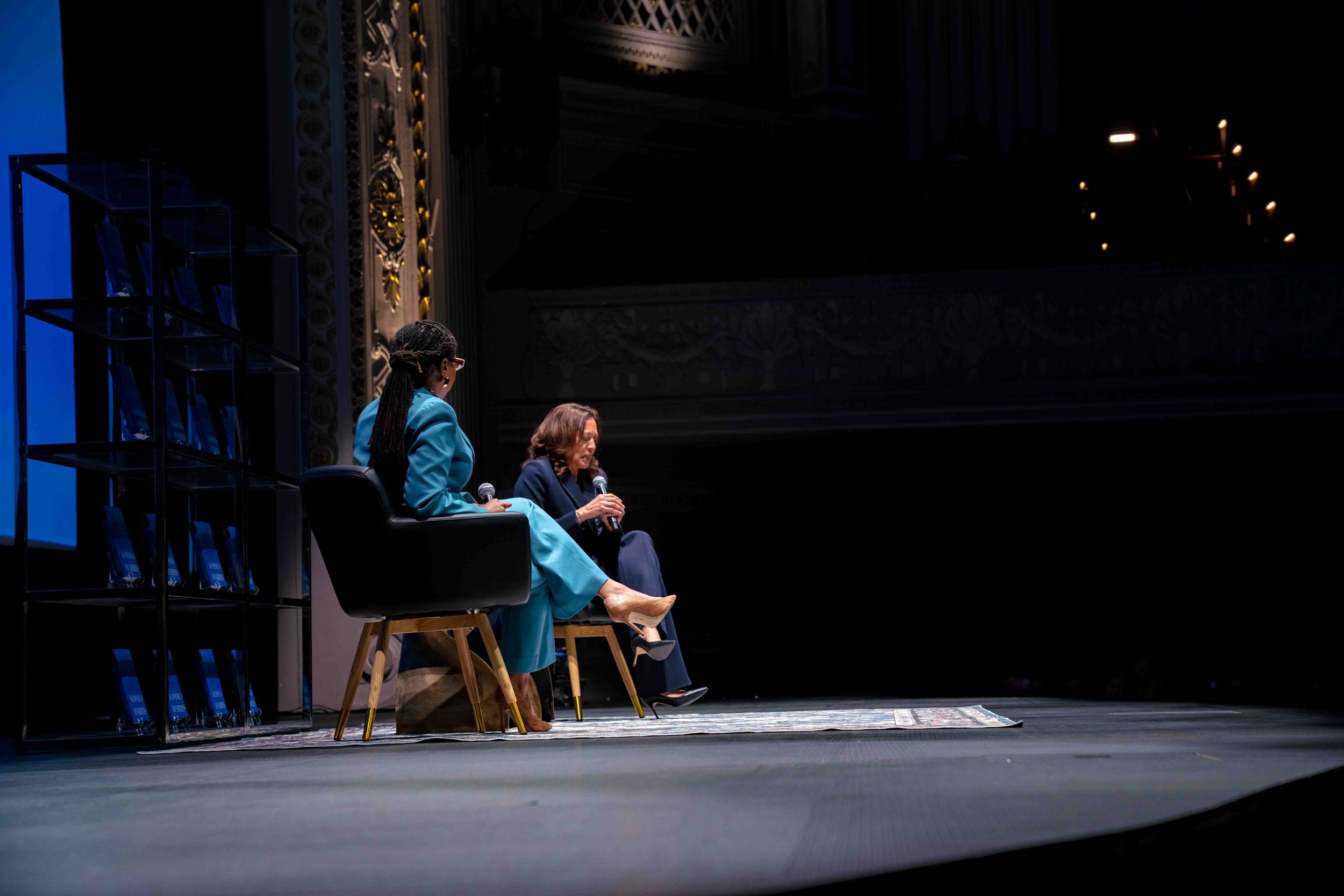 Two women sit on a stage, engaging in a discussion. One woman, dressed in a blue suit, is speaking into a microphone. The other woman, in a darker outfit, is also holding a microphone. The stage has a shelf with books or awards on the left and is dec