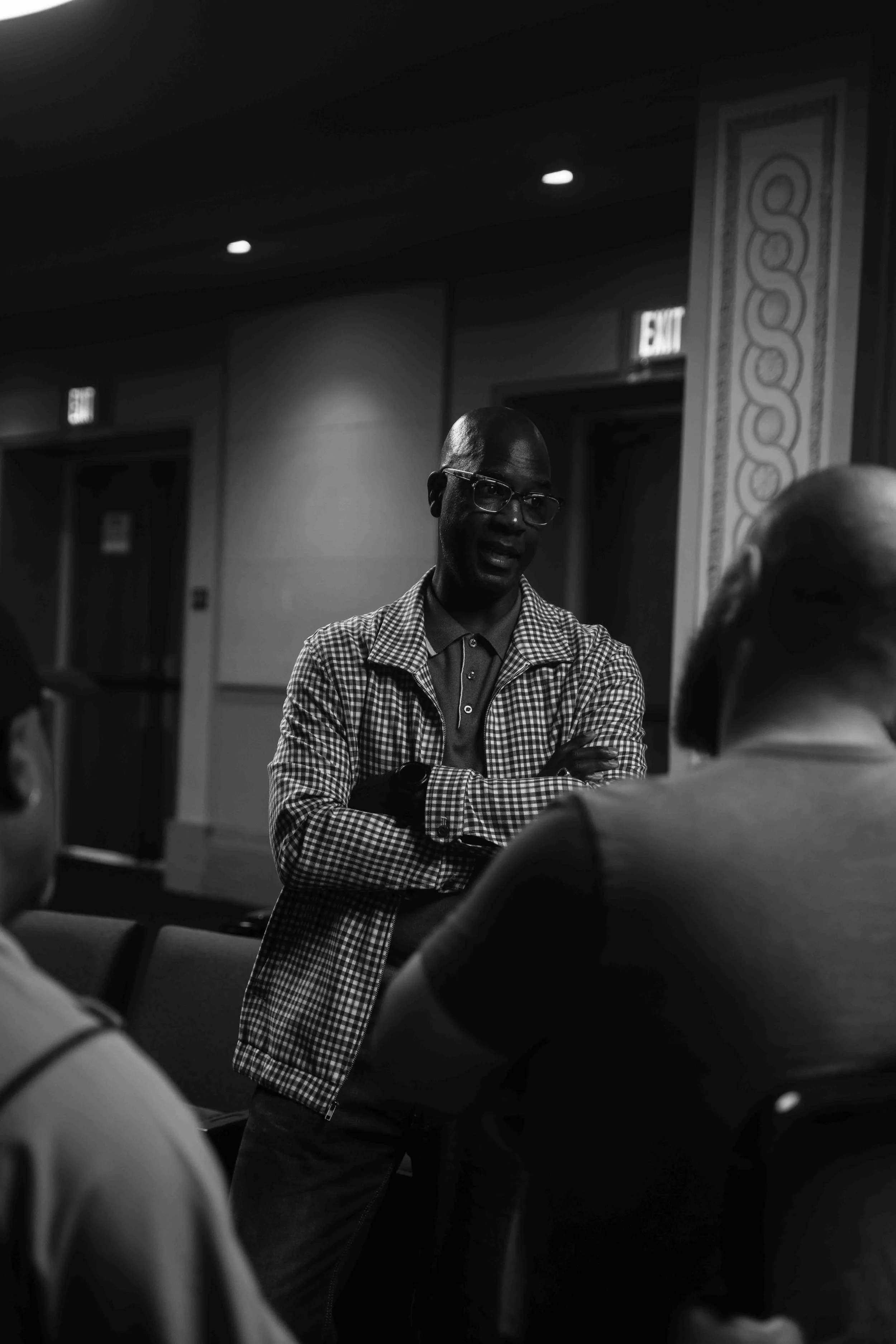 A man wearing glasses and a checkered jacket standing with arms crossed, speaking to a group in a dimly lit room with exit signs and decorative wall patterns.