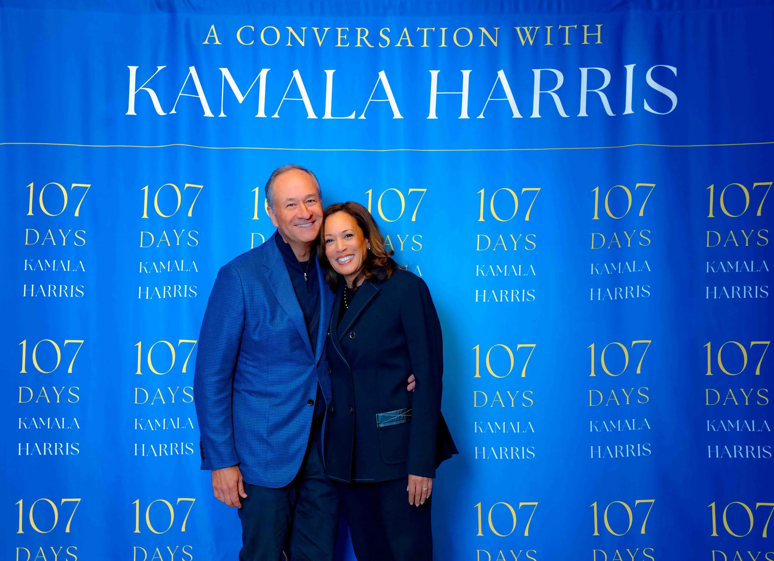 Two people smiling and standing close together in front of a blue backdrop that says 'A Conversation with Kamala Harris' and '107 Days'.