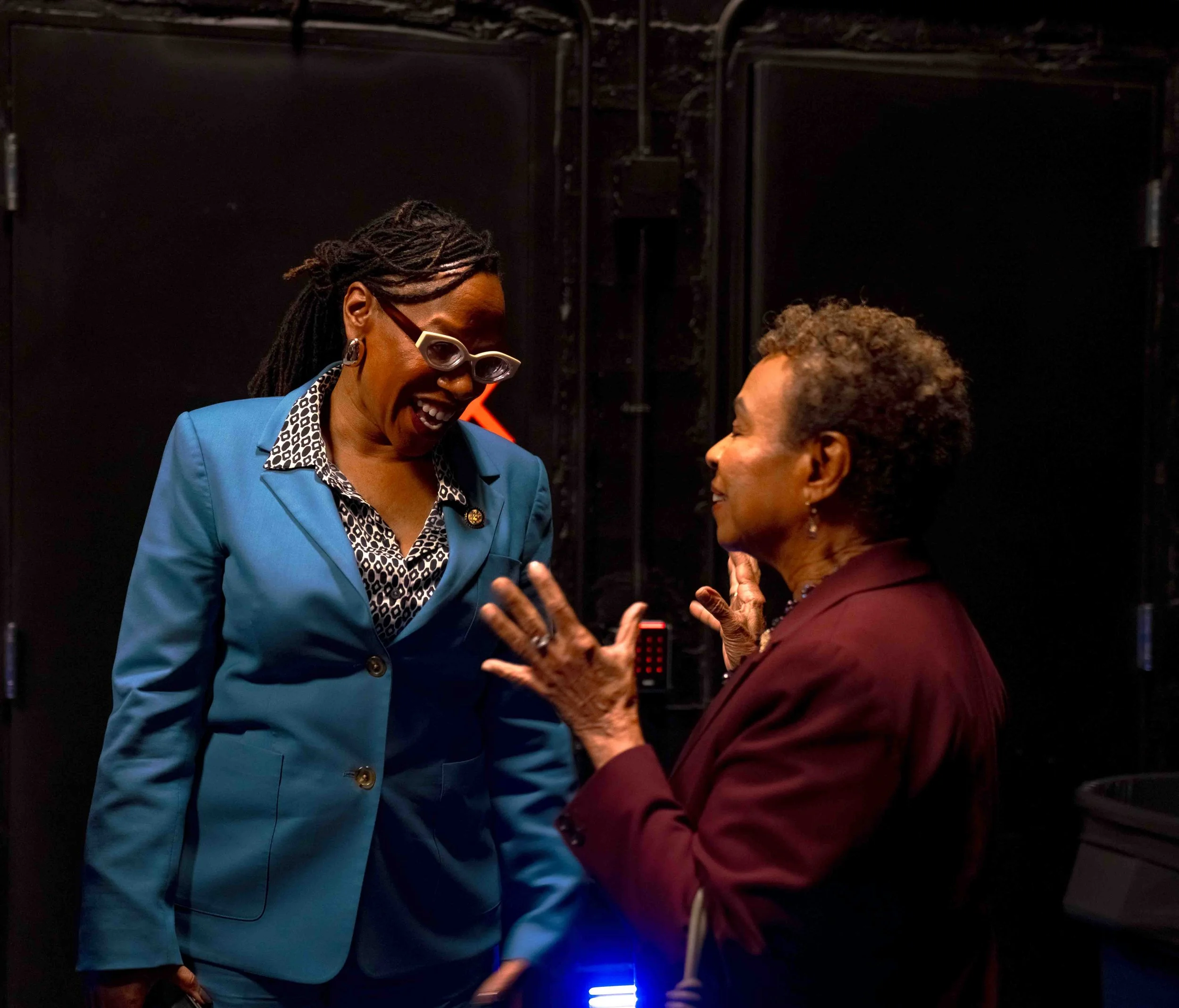 Two women are engaged in a lively conversation, smiling and gesturing with their hands. One woman wears a blue blazer and glasses, while the other wears a maroon blazer. They are indoors, against a dark background.