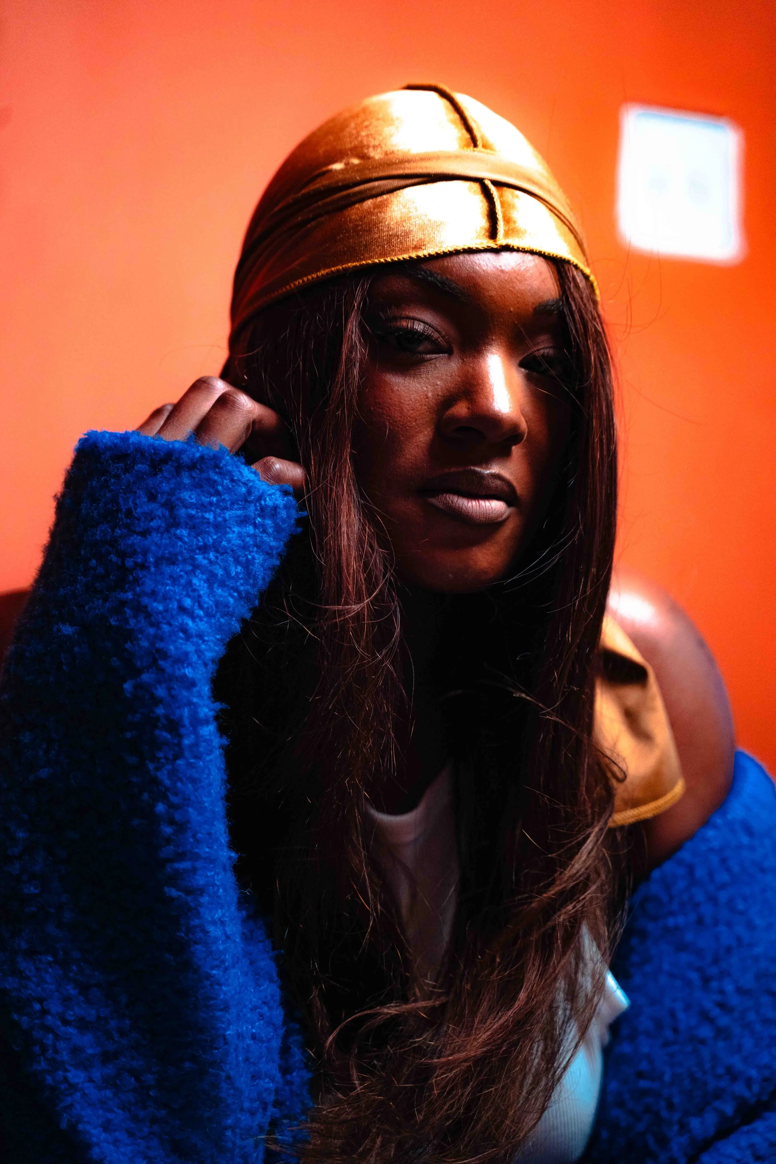 A woman with long hair wearing a gold headwrap, a blue fuzzy sweater, and a tan jacket over a white shirt, posing against an orange background.