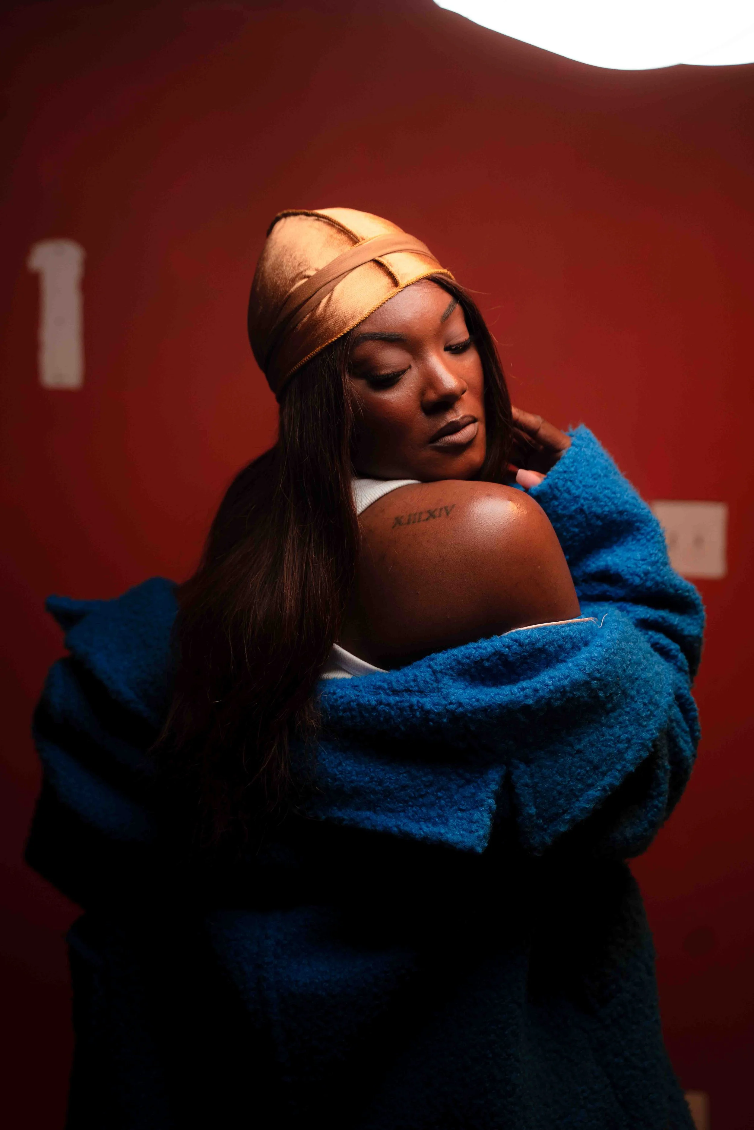 A woman with long dark hair, wearing a gold headscarf, white top, and blue coat, sitting against a red background, looking down with a contemplative expression. She has a tattoo on her shoulder.