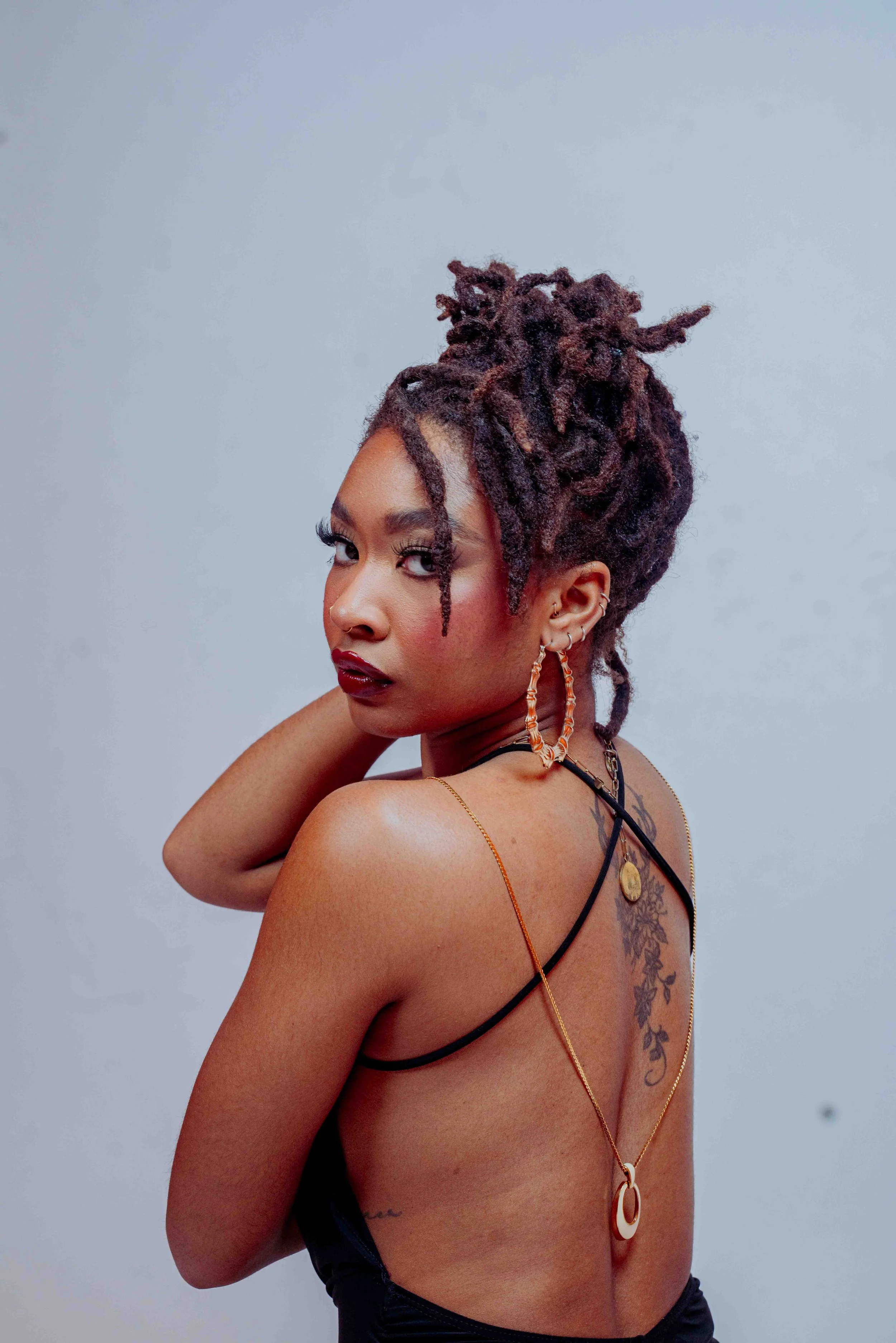 Portrait of a woman with dreadlocks, wearing hoop earrings and layered necklaces, looking over her shoulder against a plain background.