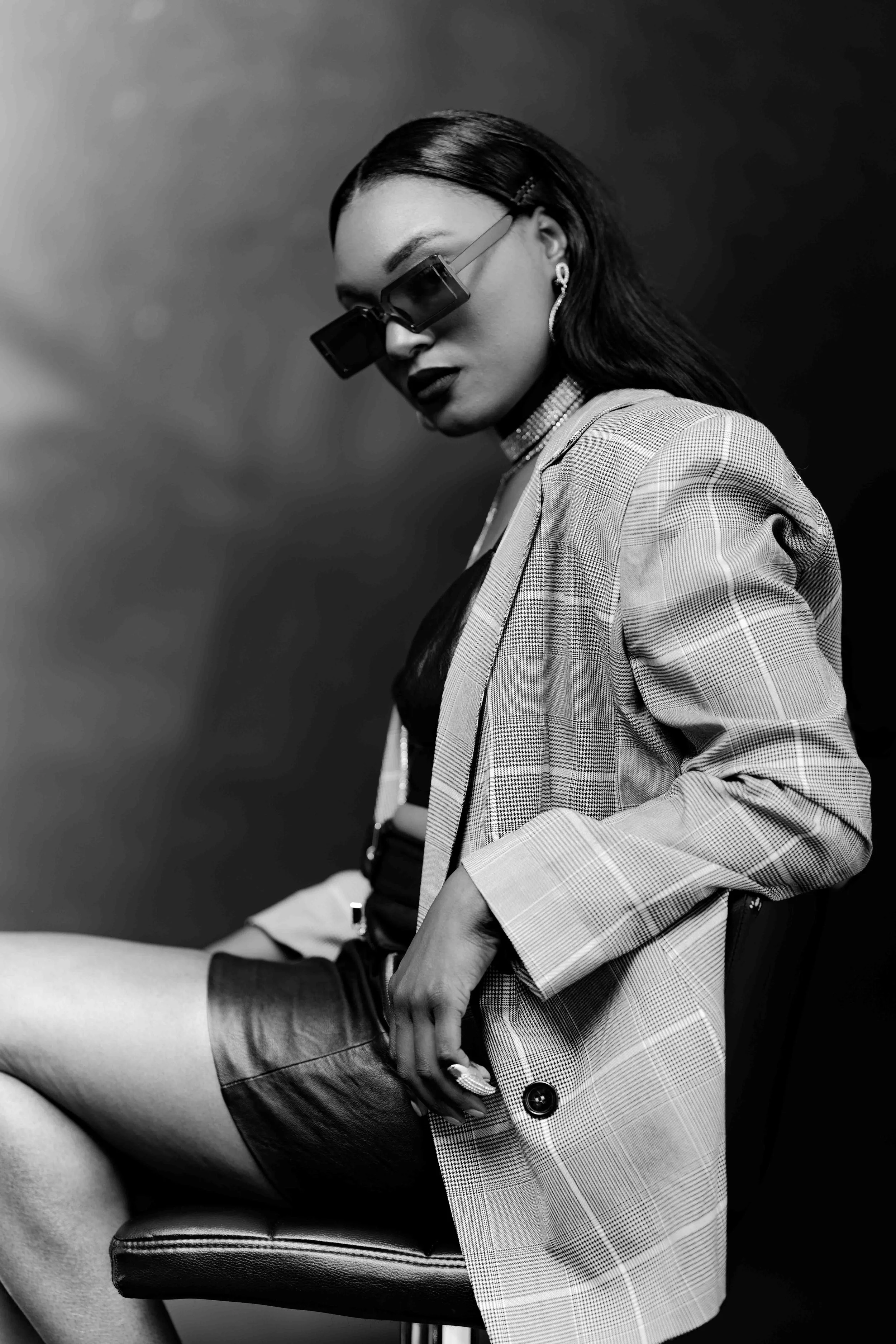 A black and white photo of a woman sitting on a chair, wearing sunglasses, a plaid blazer, a choker necklace, earrings, and a leather skirt.