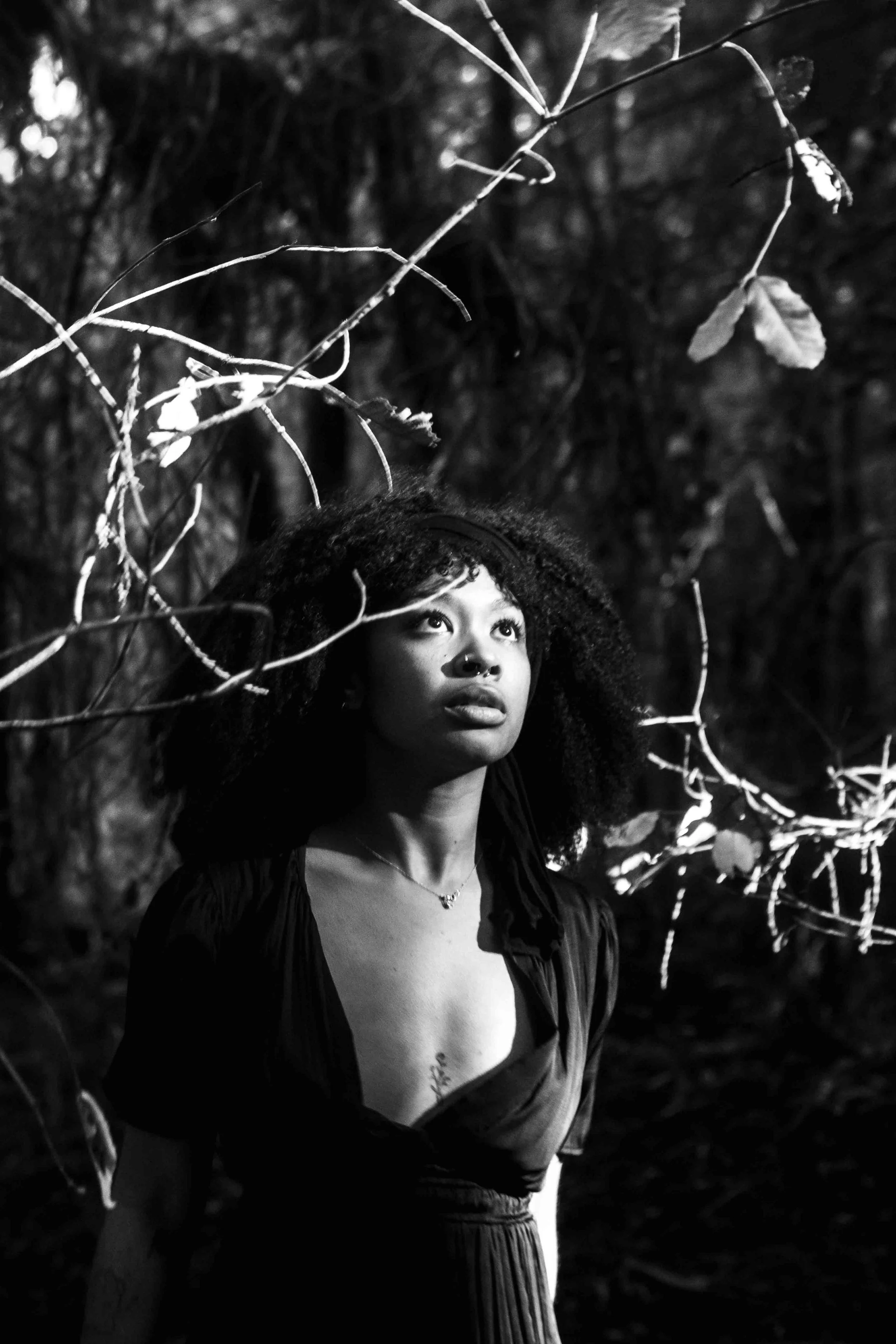A woman with curly hair and a septum piercing stands outdoors in a forest, looking up. She is wearing a dark top and has a necklace. The image is black and white and features branches and leaves around her.