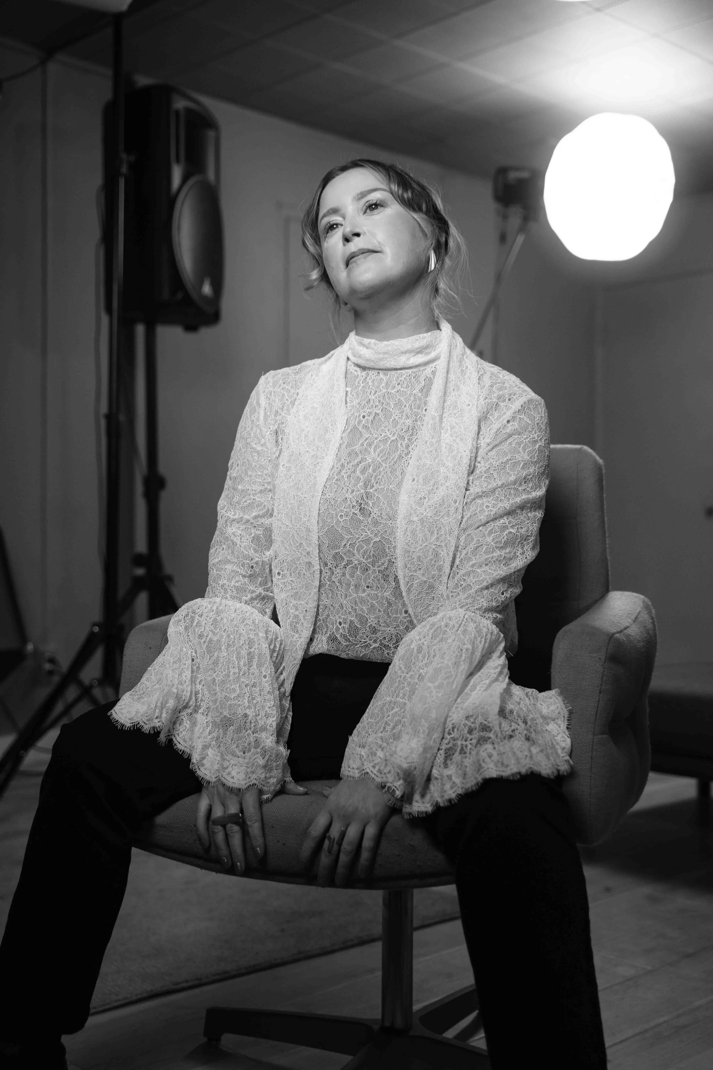 A woman with short wavy hair sits on a modern chair wearing a lace top and blazer, in front of studio lights and equipment, in a black and white photo.