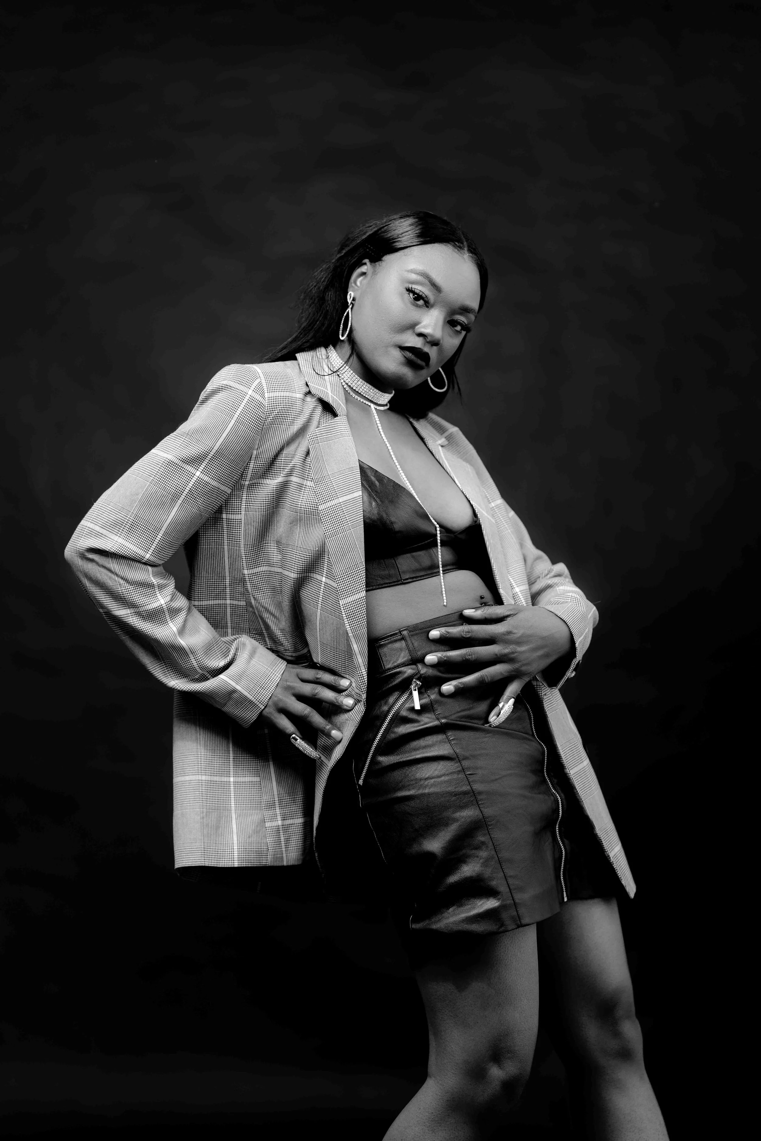 Stylish woman in leather top, leather skirt, and plaid blazer posing against a dark background in a black-and-white portrait.