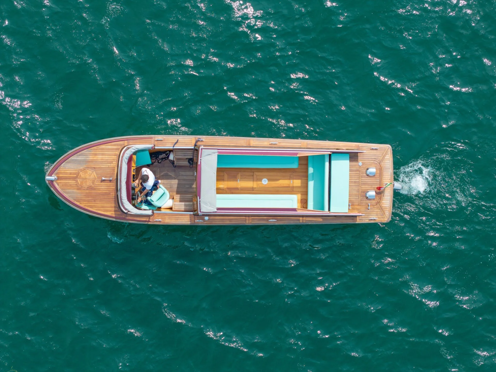 An aerial view of a wooden boat on the water with two people aboard, and the boat has teal-colored upholstery and a sunroof.