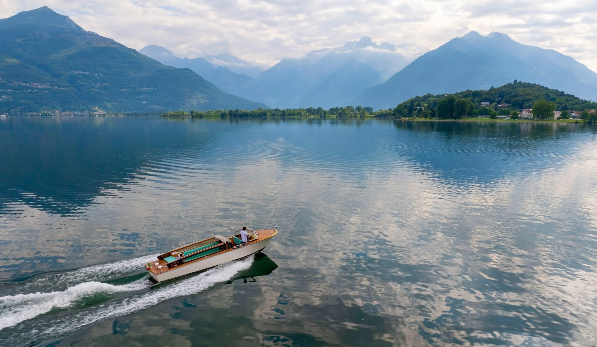 A boat cruising on a calm lake surrounded by green mountains and a cloudy sky.