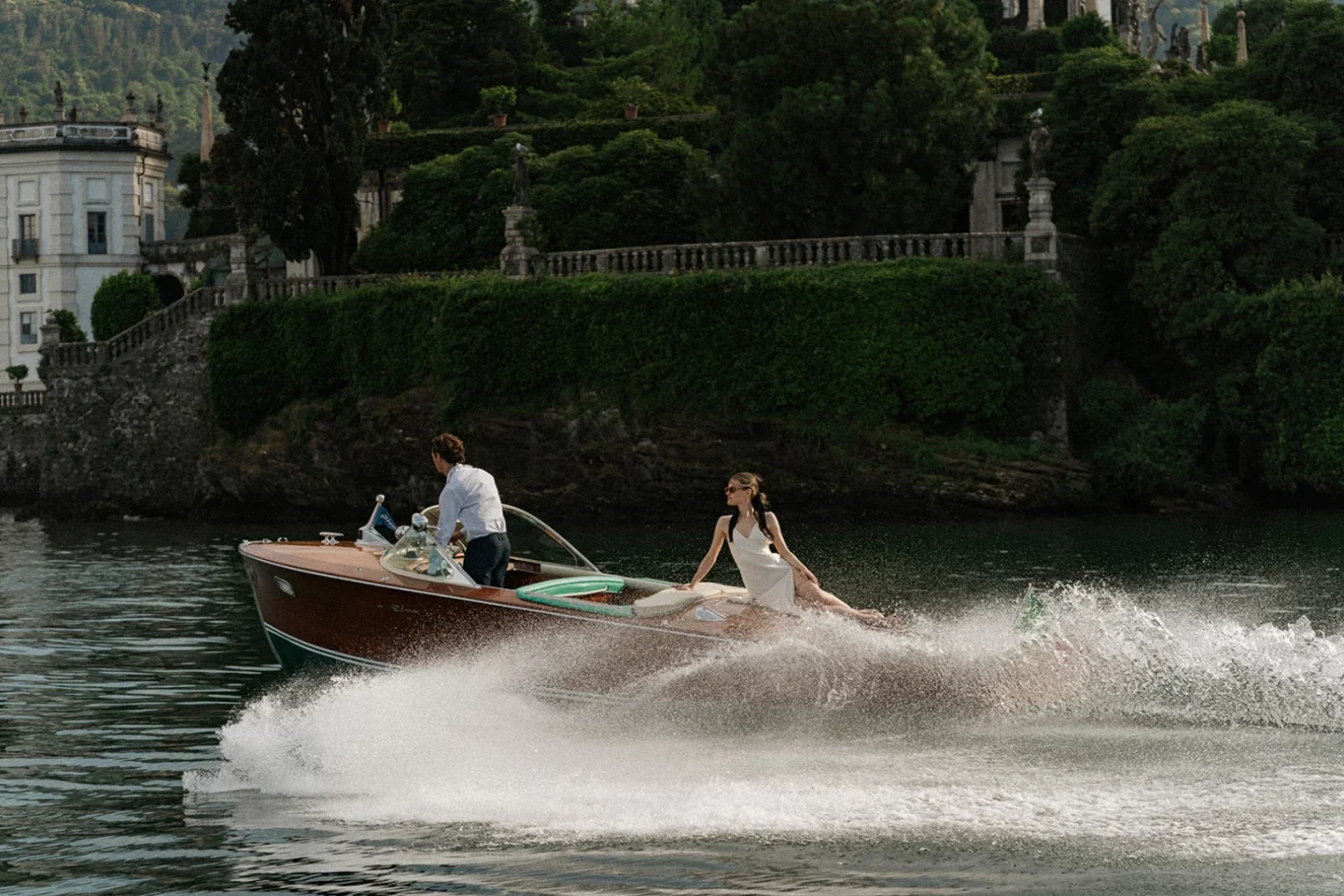 A woman in a white dress is riding on a motorboat with a man steering on a lake, with lush greenery and large historic buildings in the background.