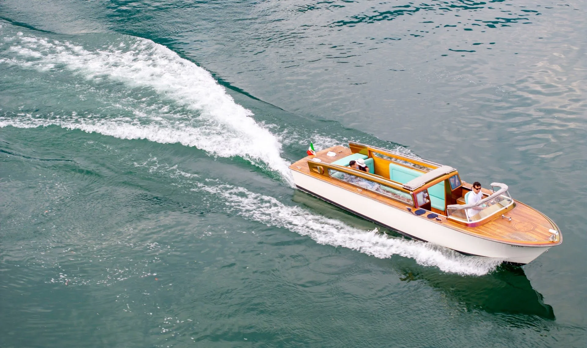 A small wooden speedboat with two passengers, speeding on a body of water, creating a wake behind it.