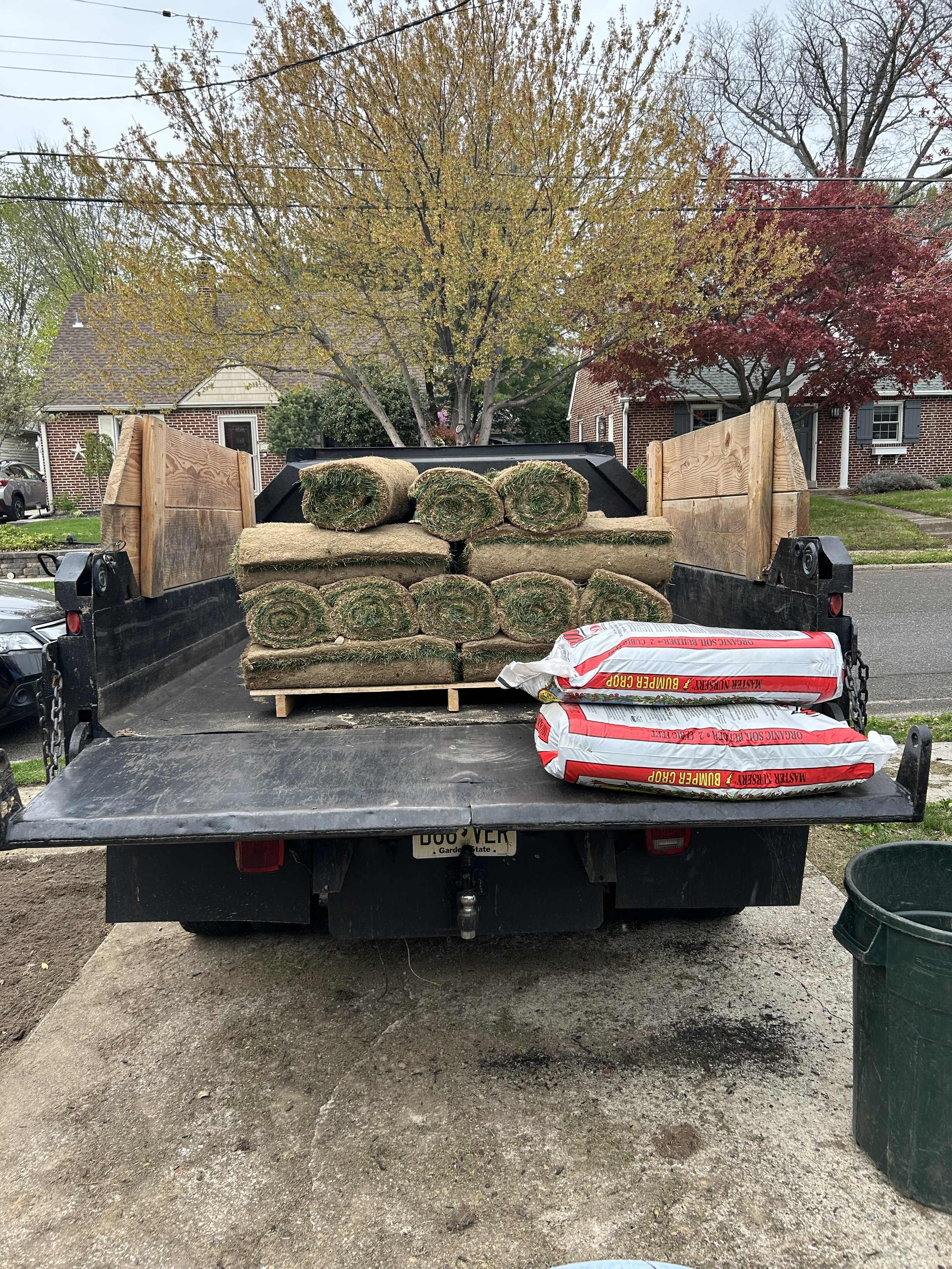 Loaded truck bed with rolls of sod, bags of mulch, in a residential neighborhood with trees and houses in the background.
