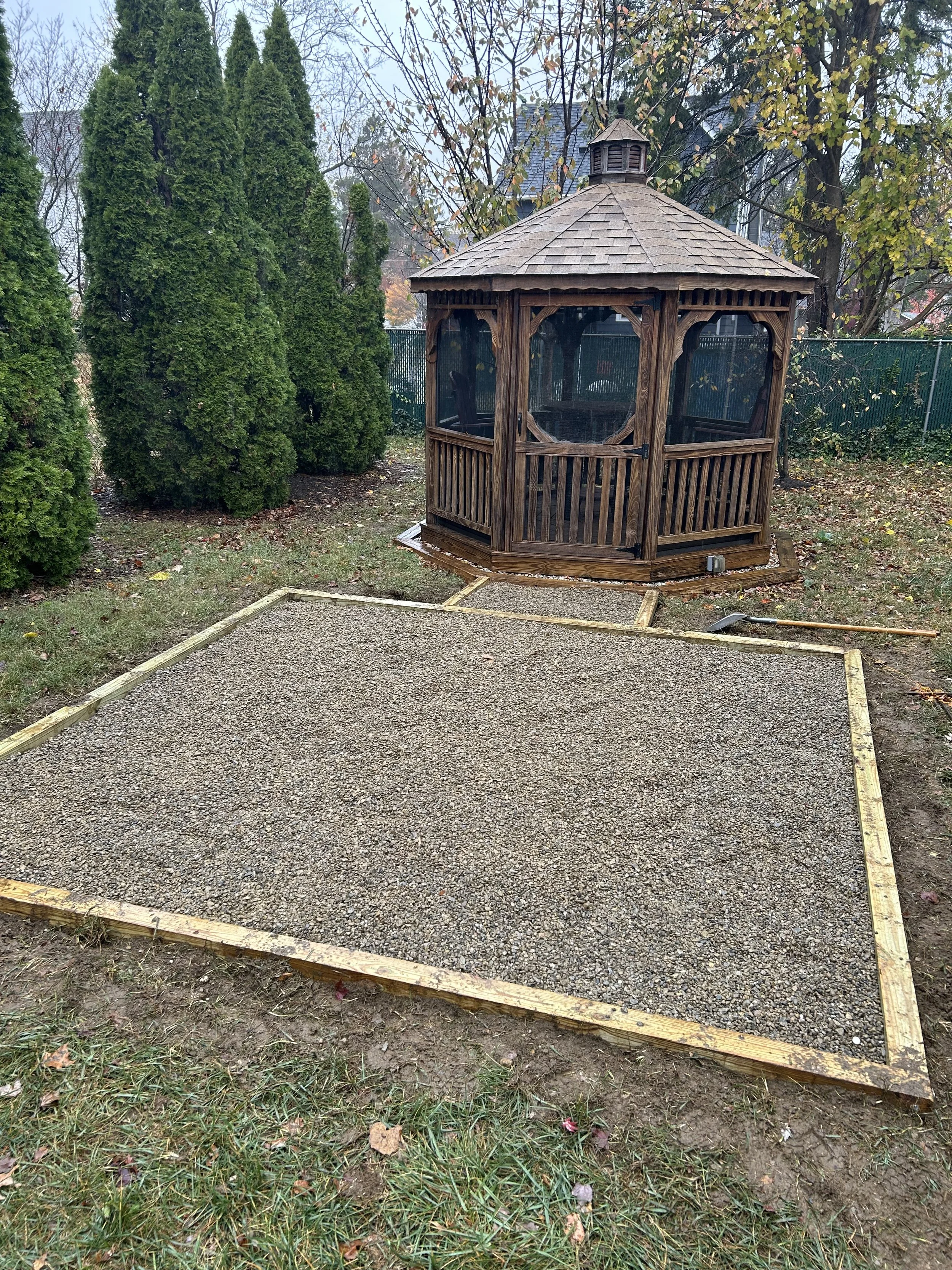 A newly built backyard sandbox with a wooden frame and gravel surface, next to a small wooden gazebo with screened walls, trees, and a fence in the background.