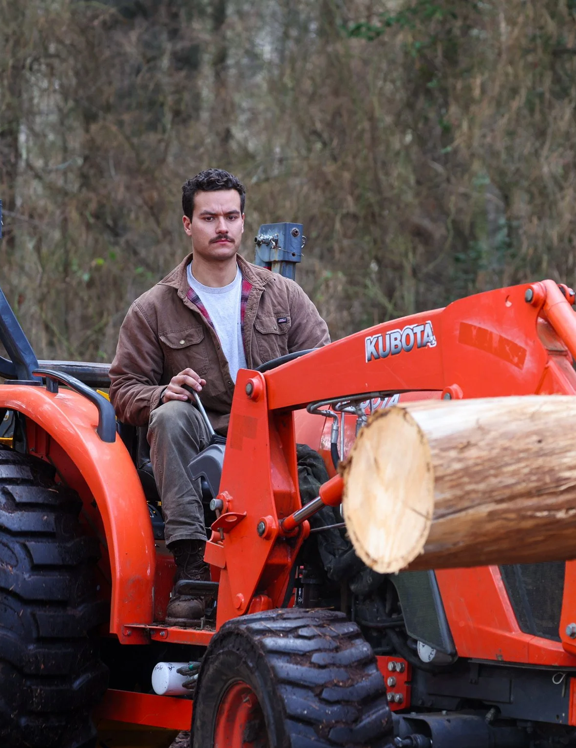 A man with dark hair, a mustache, and wearing a brown jacket and gray shirt, sitting on an orange Kubota tractor, holding a control handle, with a background of trees and a fallen log.