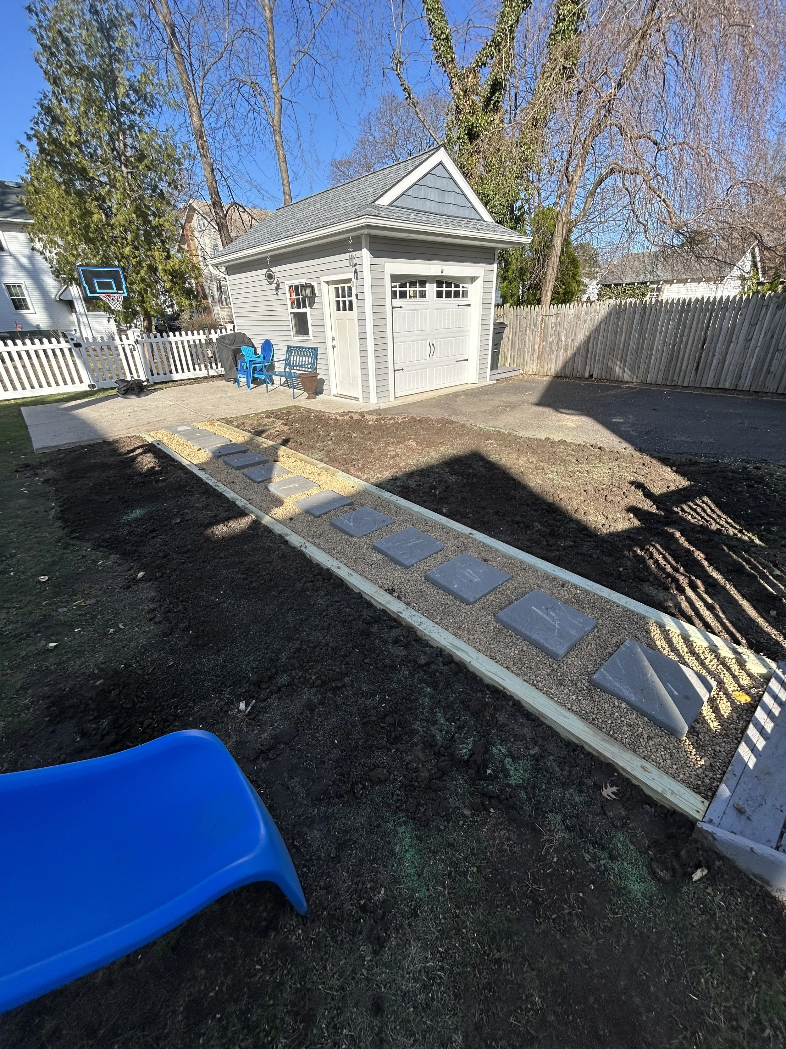 Backyard with a small building, a paved path with stepping stones, a blue slide, outdoor chairs, a basketball hoop, and a wooden fence.
