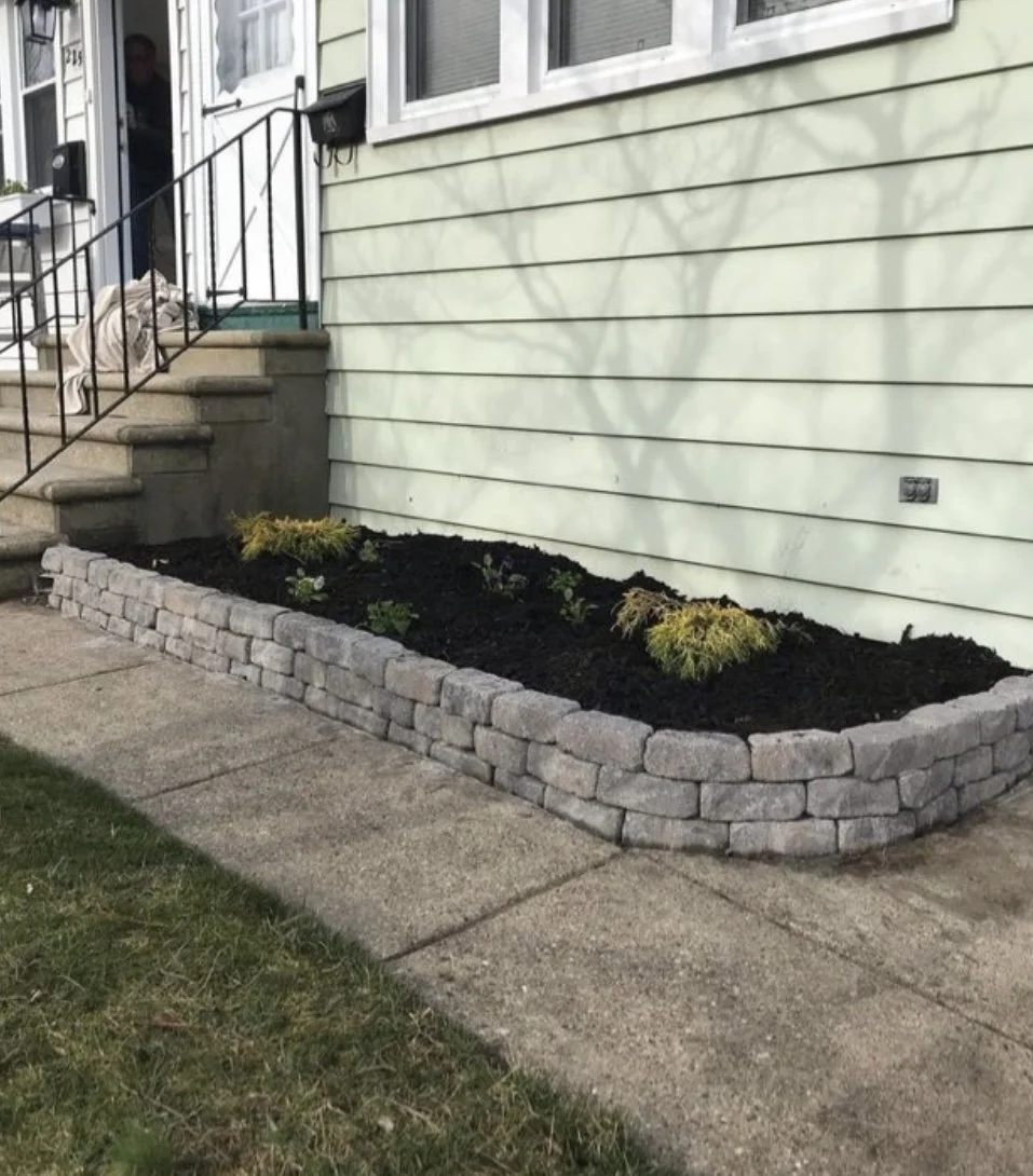 A small garden bed with young plants and small bushes, bordered by gray stone bricks, located in front of a house with light green horizontal siding. The garden is adjacent to a concrete sidewalk, with stairs and a person visible in the background.