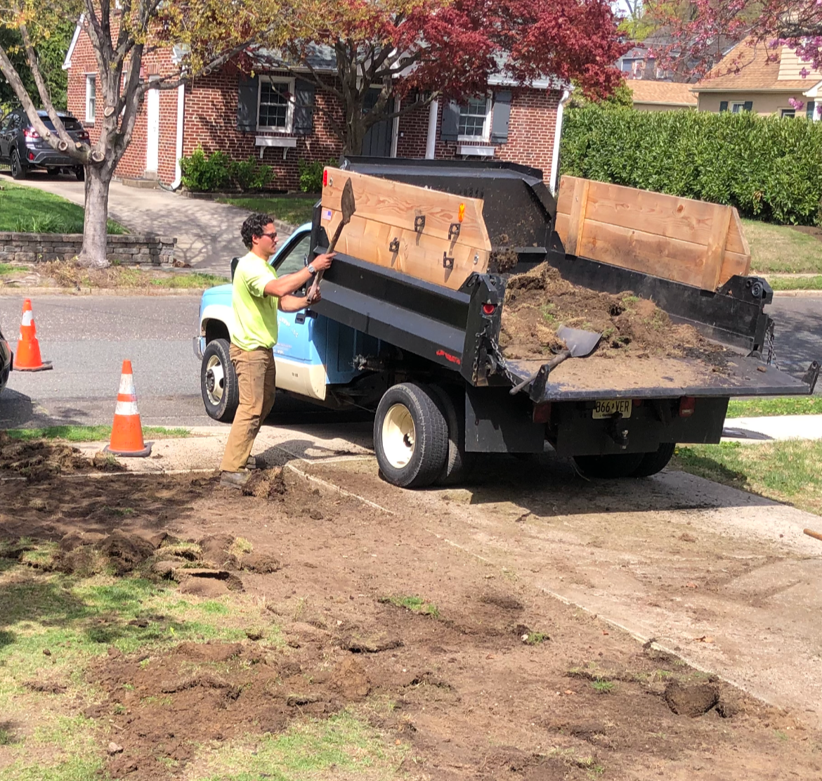 A man shoveling dirt next to a truck with a raised bed, surrounded by orange traffic cones in a residential area.